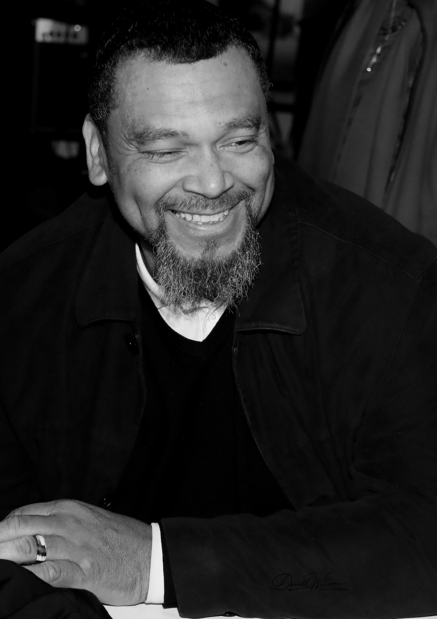 Black-and-white portrait of a smiling man with a beard and short hair, eyes closed, wearing a dark jacket.