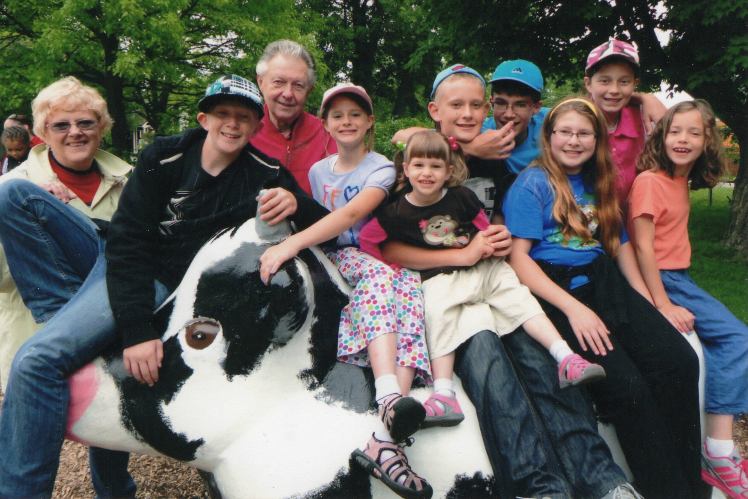 Group of children and two adults posing on a large painted cow sculpture outdoors in a park park setting, all smiling.