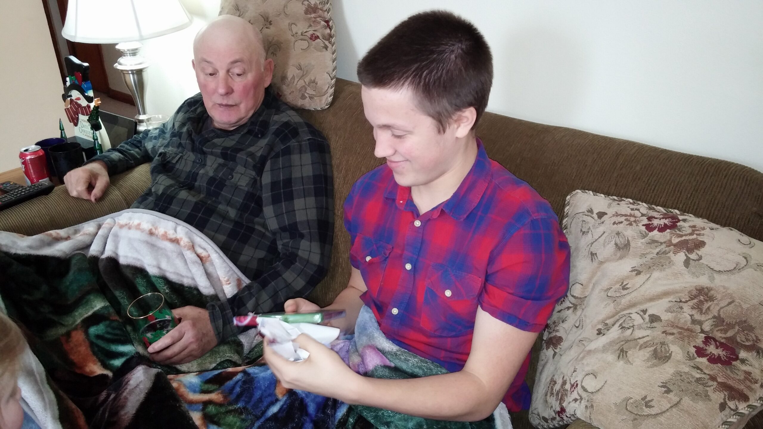 Two men sit on a brown patterned sofa indoors; the older man wears a dark plaid shirt, the younger man a red plaid shirt, both wrapped in a colorful blanket and looking at gifts or items in their laps.