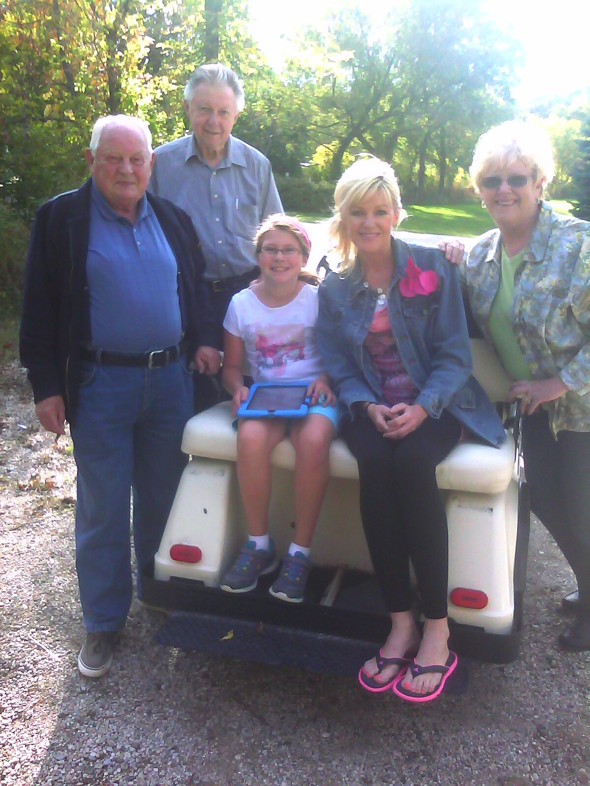 Five people pose together in a sunny park, sitting on the back of a small white utility cart with the others standing nearby.