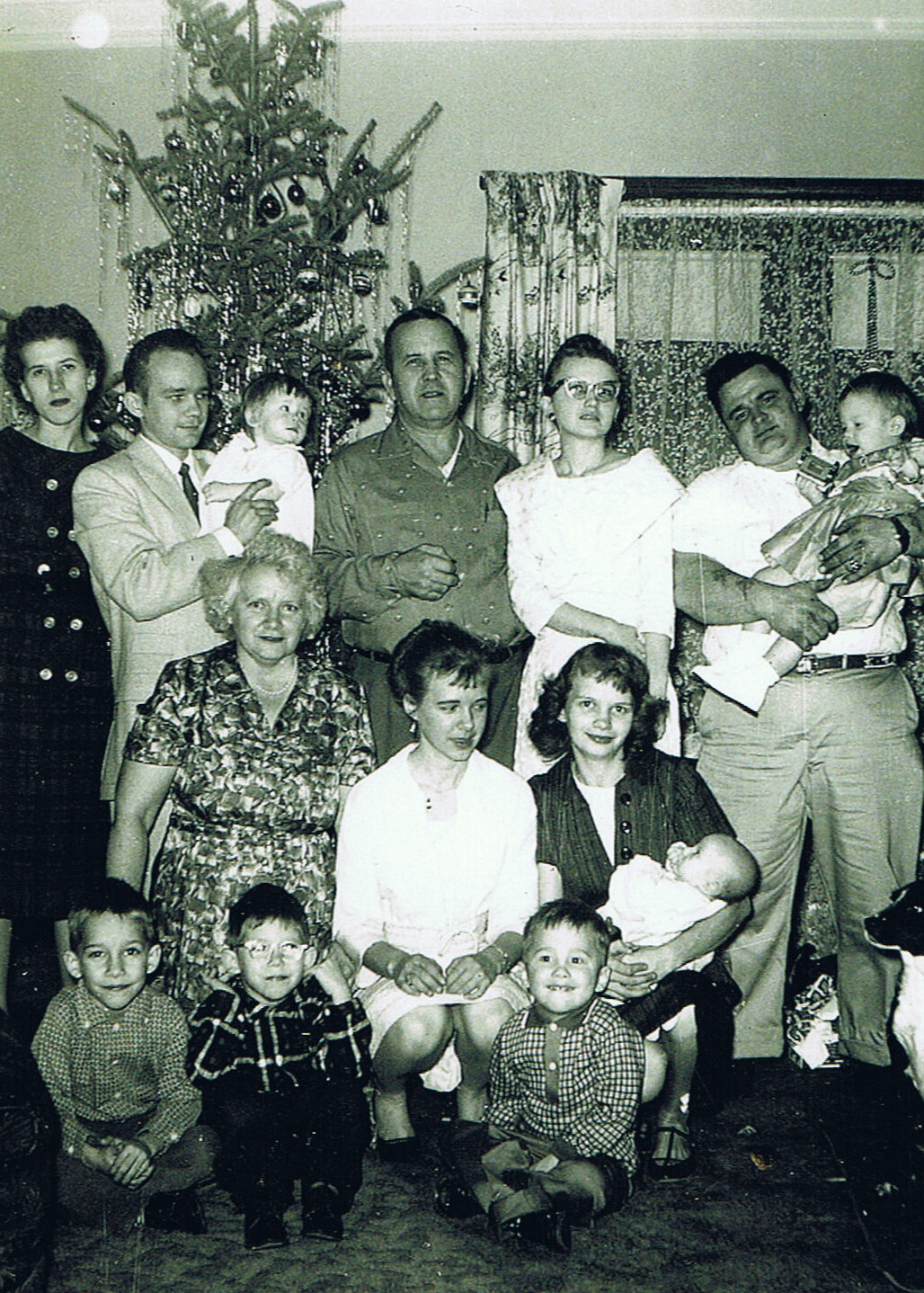 Family group posing around a decorated Christmas tree in a living room, with children in the front and adults behind, some holding babies.