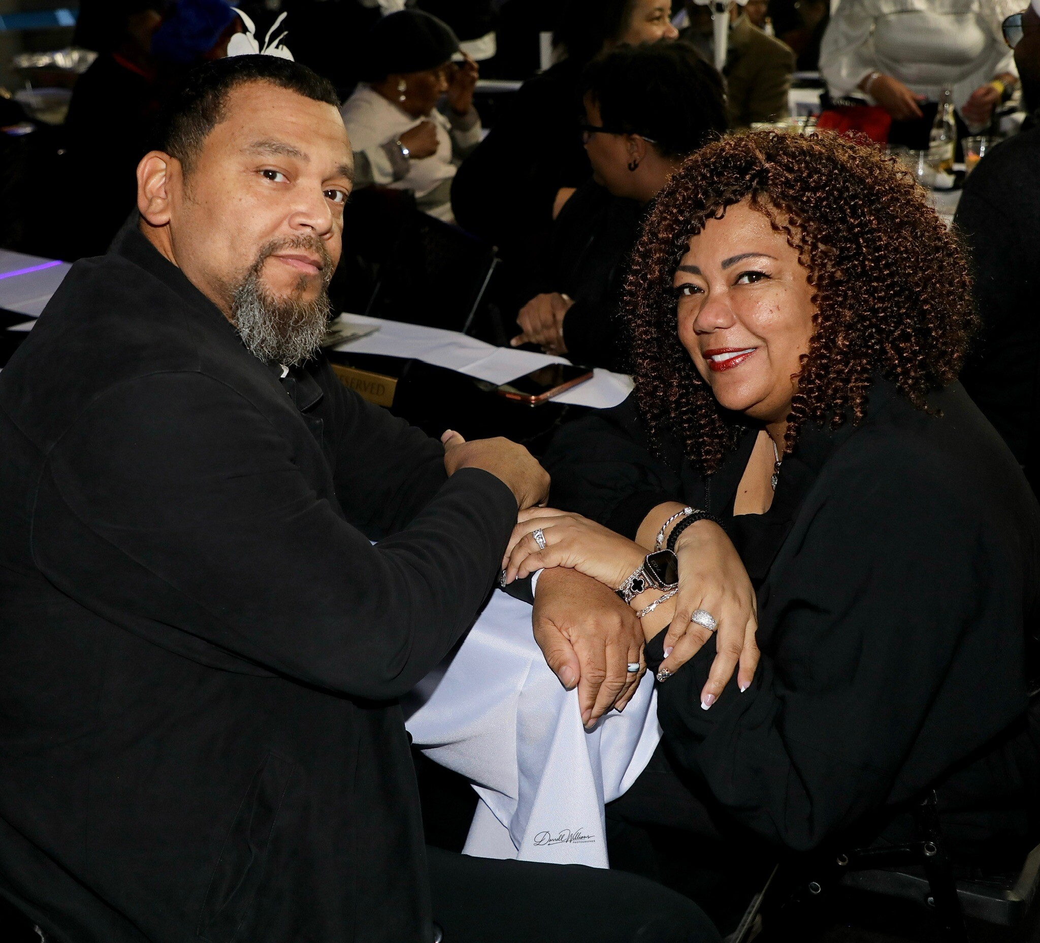 Man and woman seated at a table, holding hands and smiling at a formal social event.