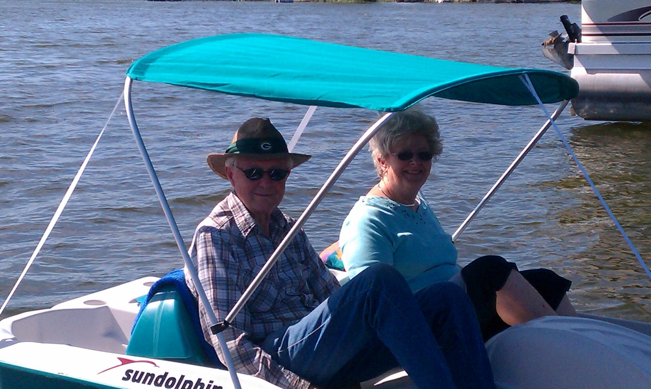 Two smiling seniors wearing sunglasses sit in a small turquoise-canopy boat on a lake. A logo reads SunDolphin on the side of the boat.