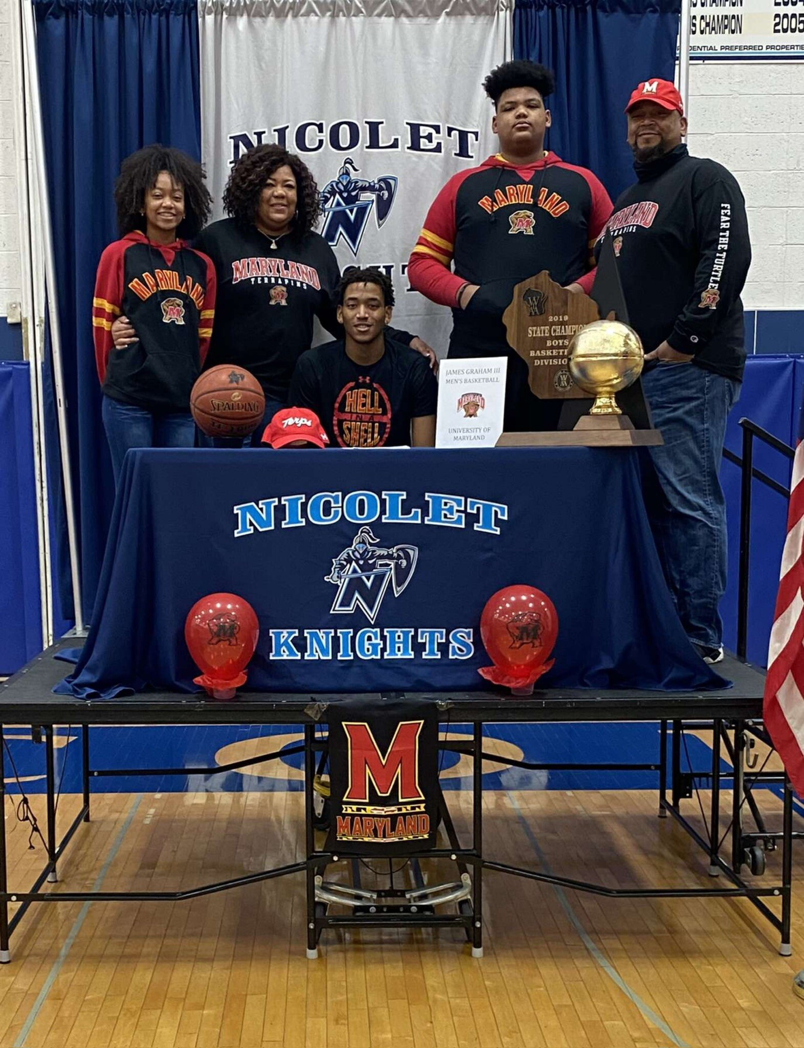 Group portrait of Maryland Knights basketball players and coach standing behind a table with a trophy and a banner that says NICOLLET KNIGHTS; a basketball, a cap, and a sign sit on the table.