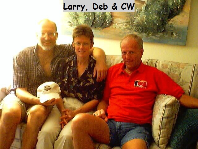 Three adults sit close together on a sofa in a living room, smiling at the camera; the man on the left holds a white cap in his lap.