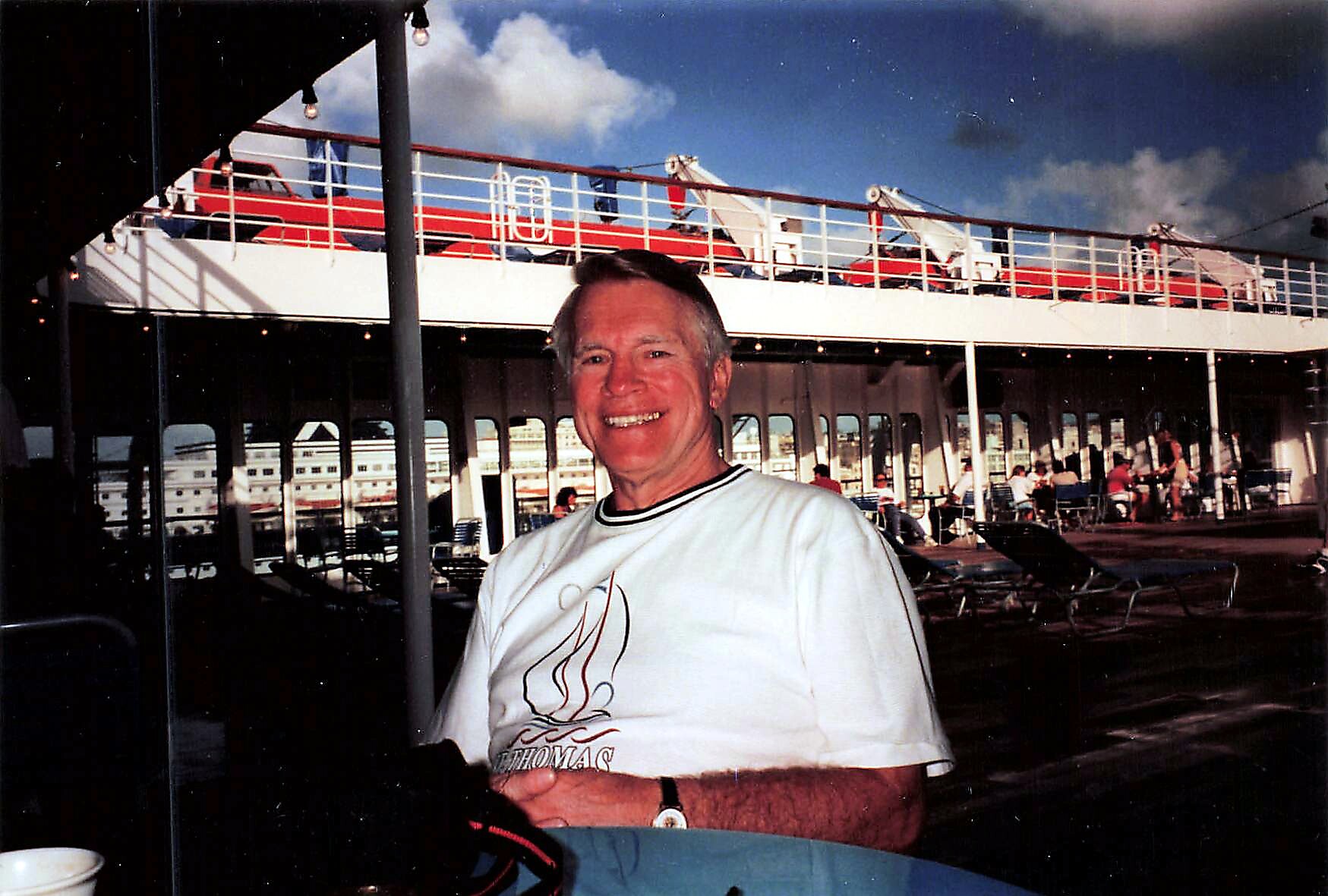 Smiling man in a white T-shirt sits on a cruise ship deck, with a blue sky and lifeboats visible above him.