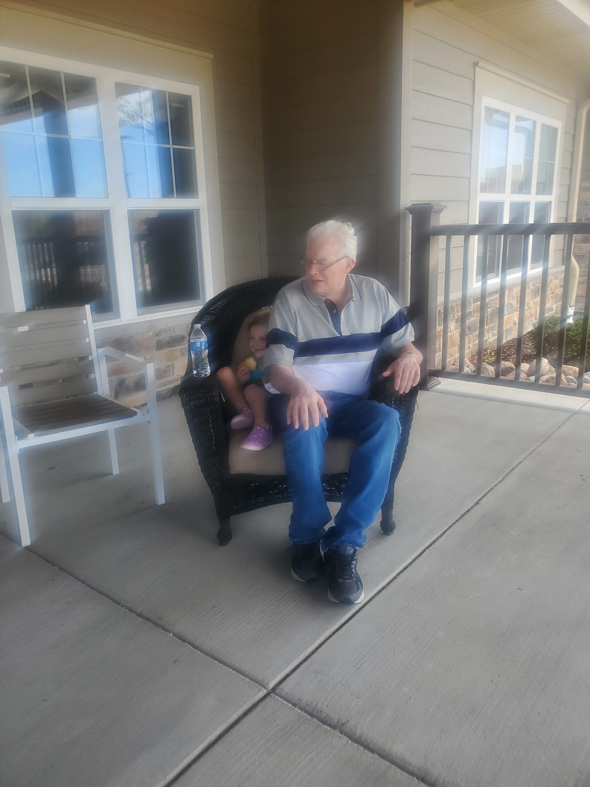 Older man in a polo shirt sits in a wicker chair on a porch, a young girl beside him, a water bottle on a nearby table.