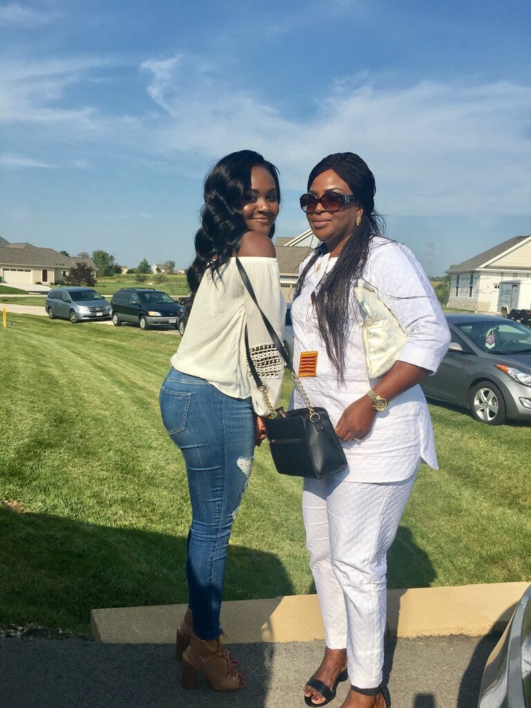 Two women posing together outside on a sunny day in a suburban park; one wears an off-the-shoulder top and jeans, the other a white outfit with sunglasses and a black handbag.