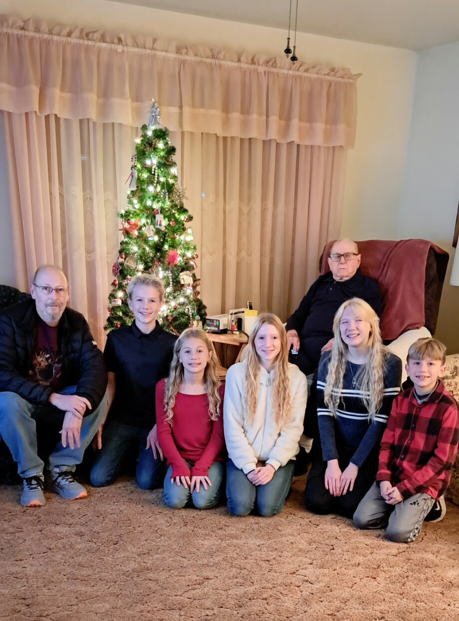 A multi-generational family posing for a Christmas photo around a lit, decorated tree in a cozy living room.