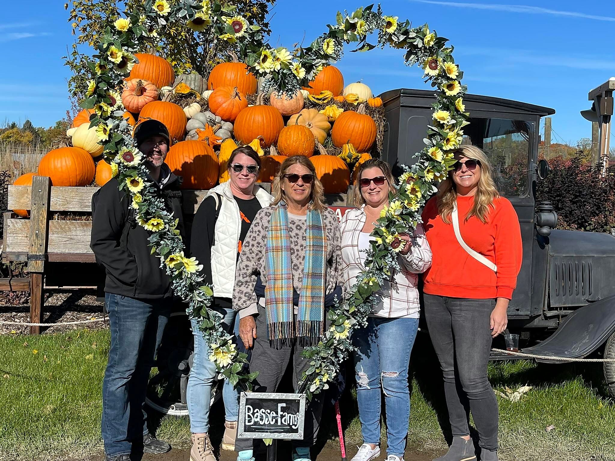 Five adults pose in front of a pumpkin-filled display on a flatbed truck, framed with a sunflower garland for a fall festival at Bassie Farms.