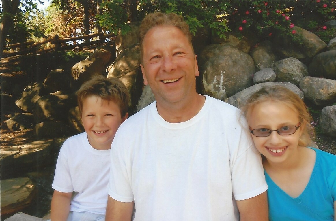 Father with two kids posing outdoors, rocky garden backdrop and greenery, all smiling at the camera.