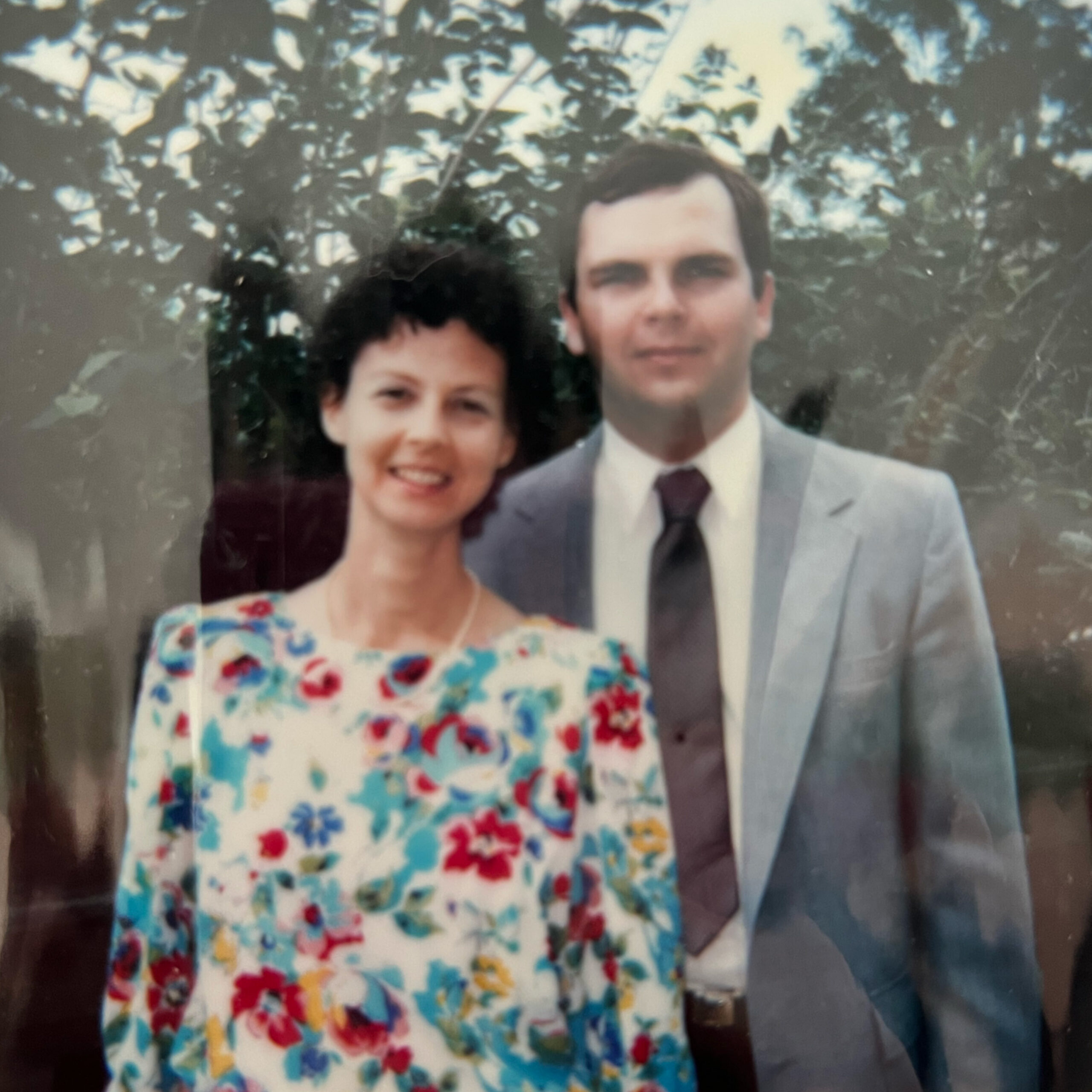 A smiling couple posing outdoors, woman in a floral dress and man in a light suit, with trees in the background (blurry vintage photo).