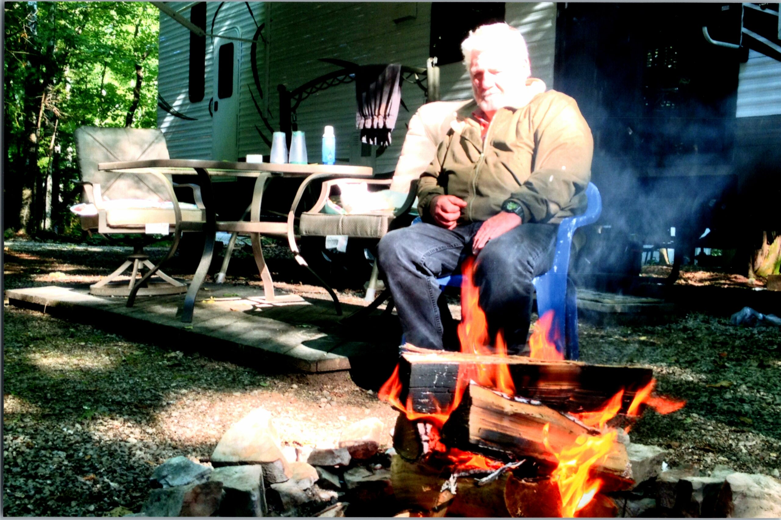 Older man in a tan jacket sitting in a blue plastic chair by a campfire outside a trailer, with a wooden table and patio chairs nearby.