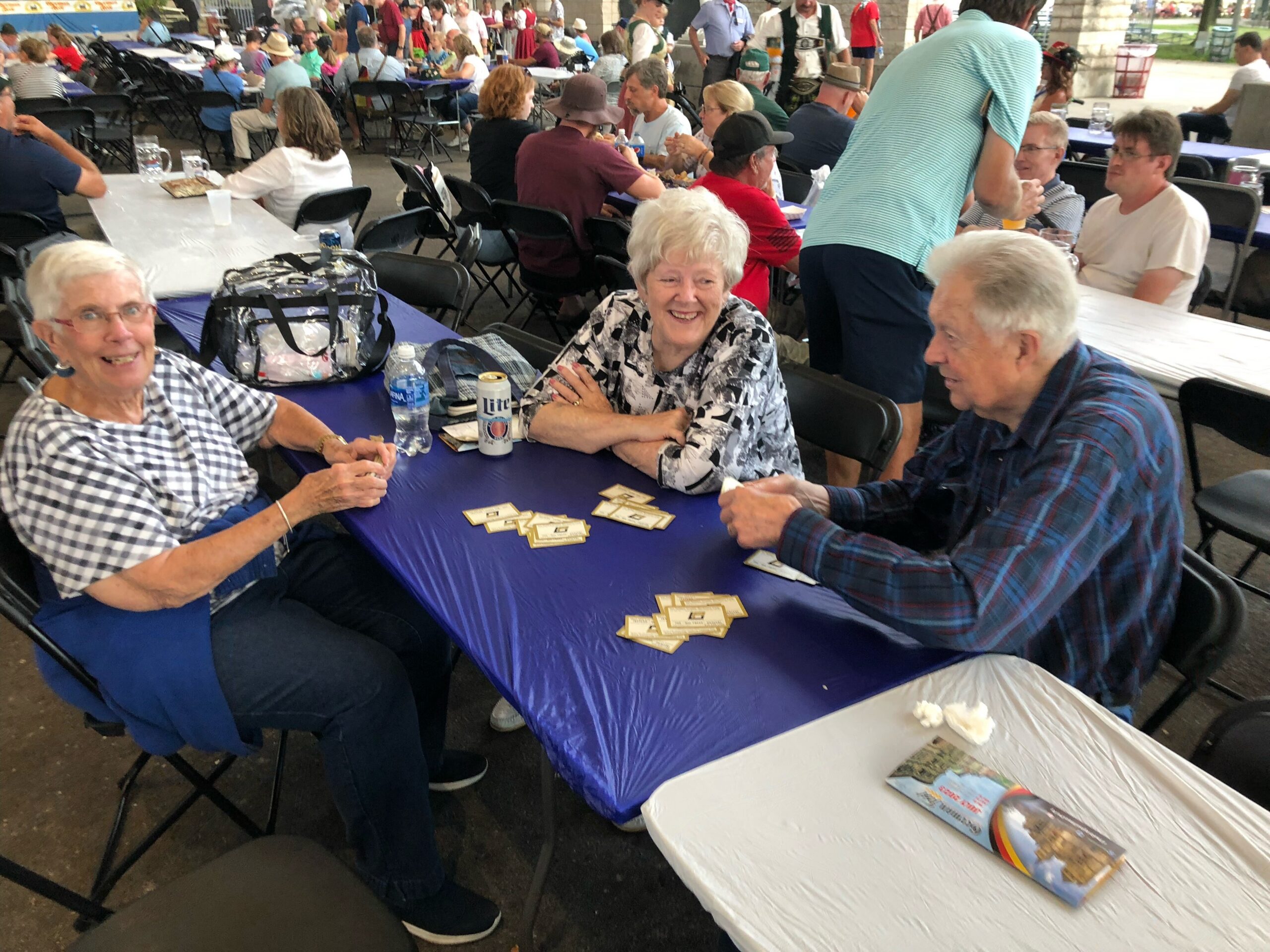 Seniors sitting at a long blue-tablecloth table outdoors, playing cards and chatting at a crowded community event.