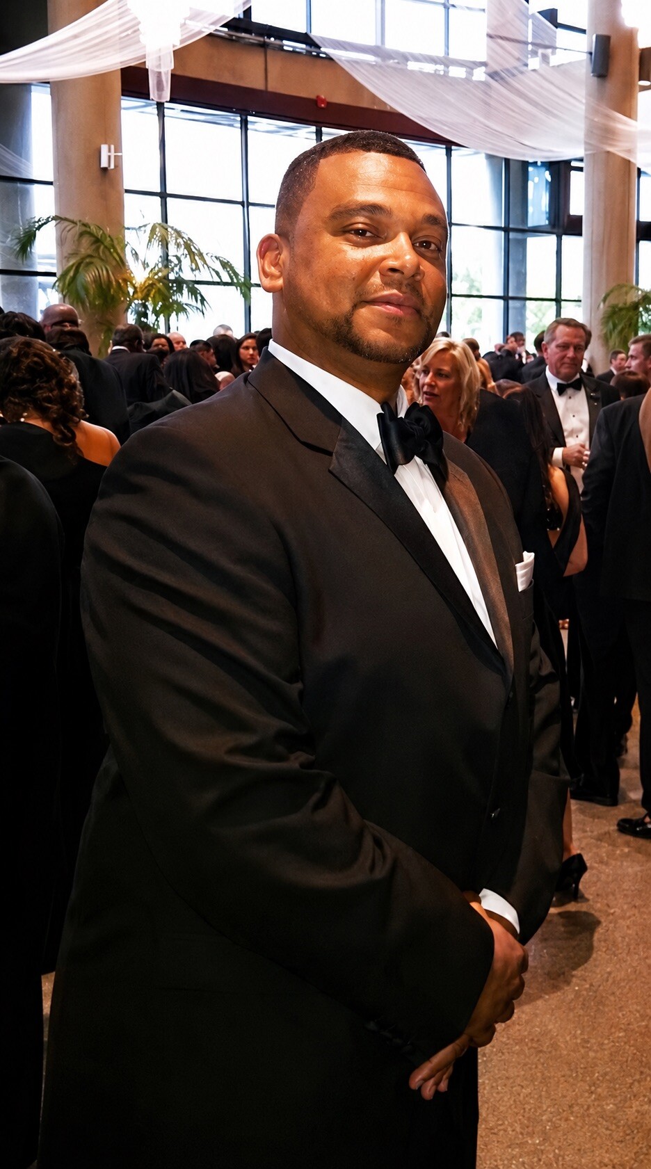African American man in a black tuxedo and bow tie posing at a formal gala, with others in evening wear in the background.