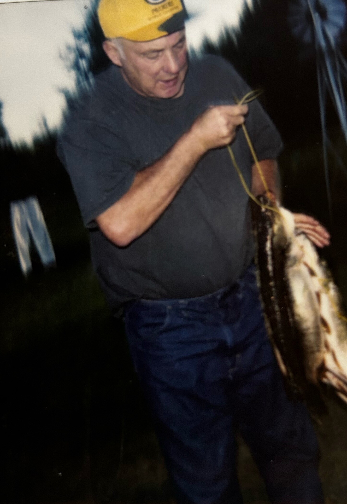 Man in a yellow cap holds a rope and a large fish outdoors at dusk, with trees in the background.