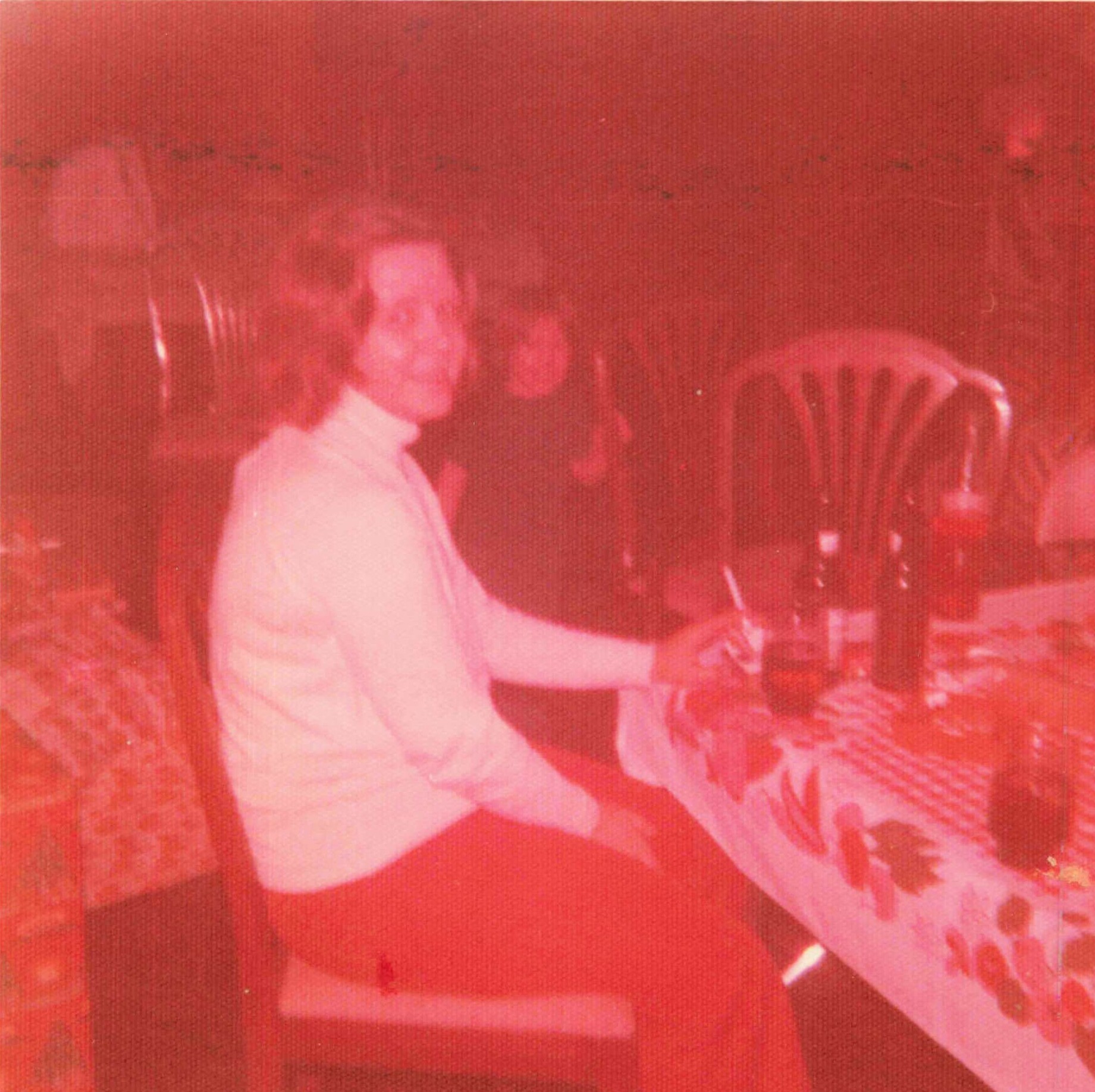 Two people at a casual indoor gathering; a man in a light shirt stands near a seated person at a table with drinks in a dim, red-tinted room.