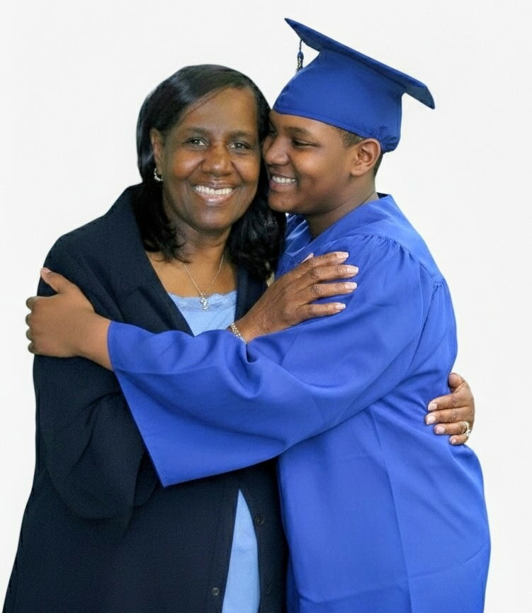 Graduate in blue cap and gown embraces a woman in a dark blazer, smiling against a white background