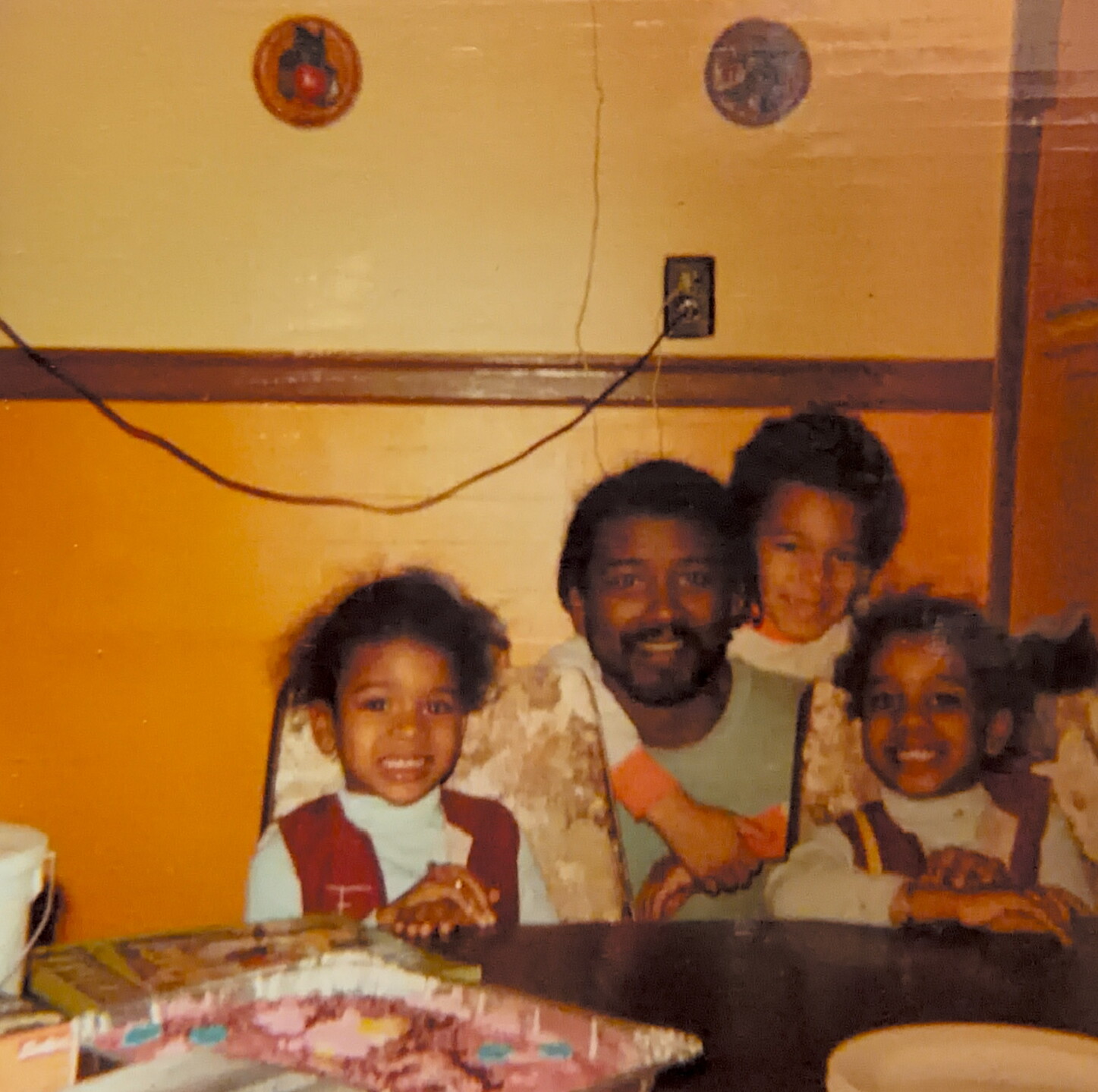 Family group photo at a birthday party: four smiling children gathered around a cake on a table, indoors against a yellow wall.