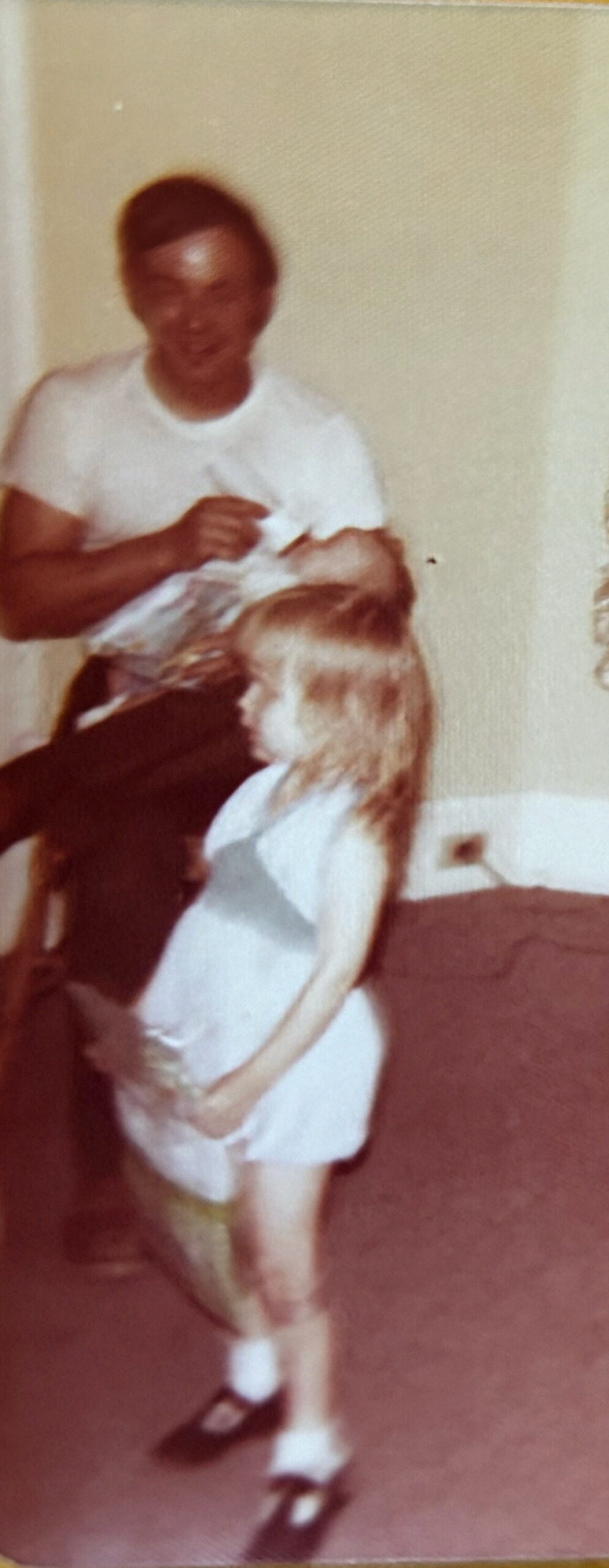 Man sits indoors in a white shirt, holding a baby wrapped in a blanket with a fluffy brown-and-white dog nearby.