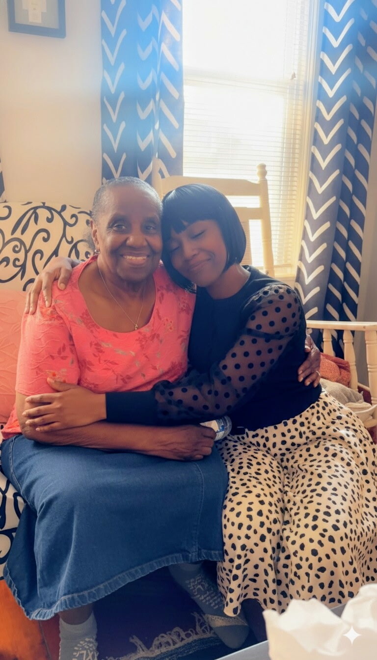 Two women sit closely on a sofa, smiling, as one hugs the other in a cozy living room.
