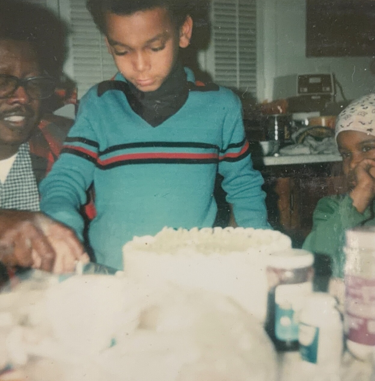 Young boy in a blue sweater cutting a white frosted birthday cake while family members watch nearby at a dining table.