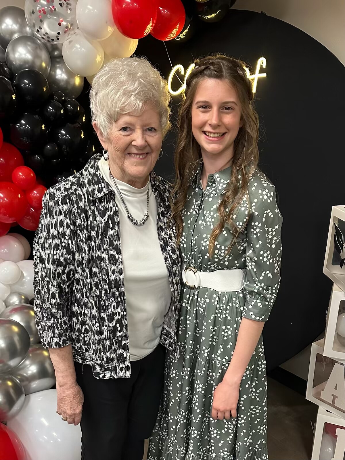 Two generations posing together at a celebration, smiling in front of a balloon arch backdrop with black, white, red balloons.