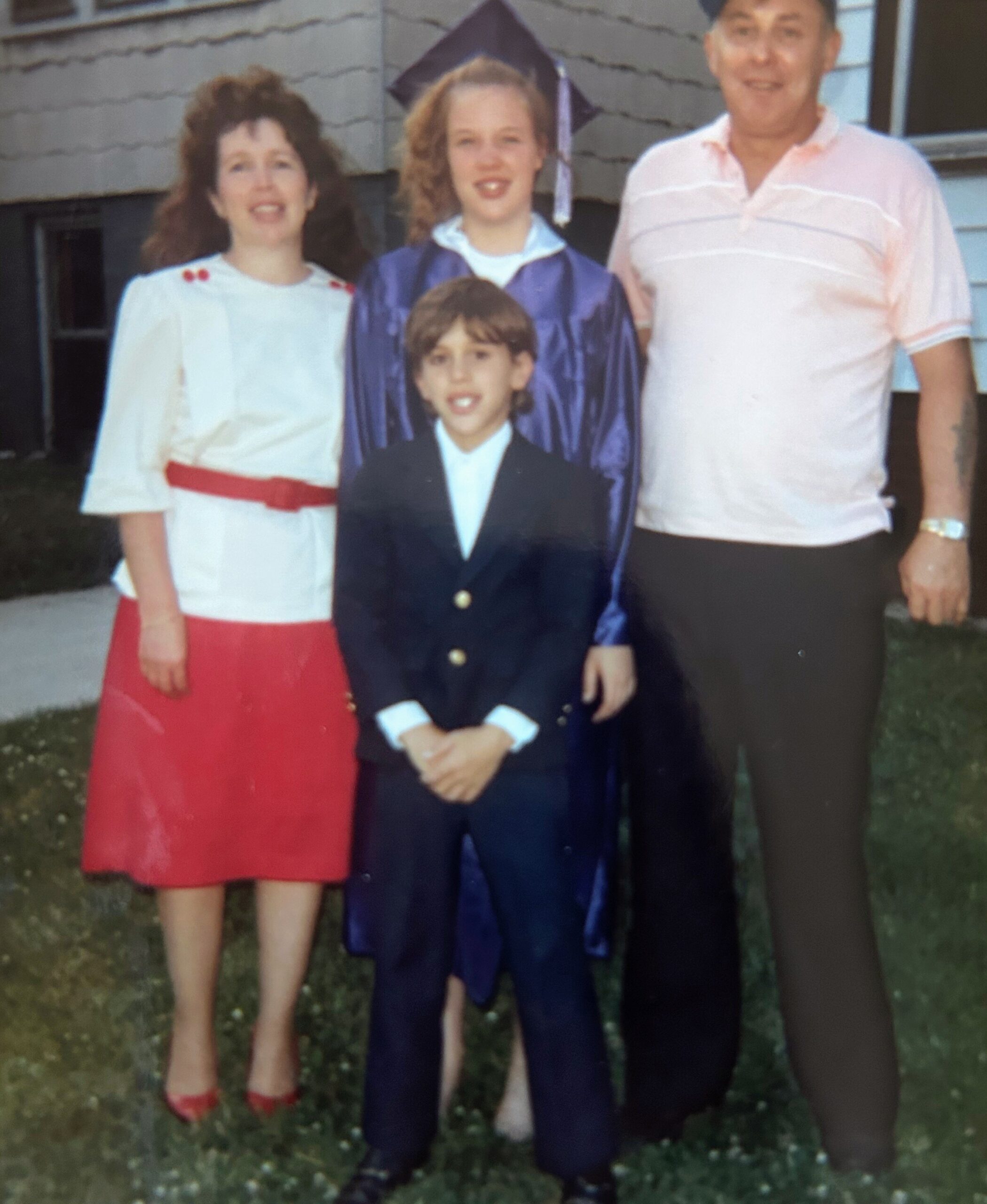 Family of four posing outside for a graduation photo: a graduate girl in a purple cap and gown stands behind a young boy in a dark suit, with a woman in a white blouse and red skirt on the left and a man in a light pink polo on the right.