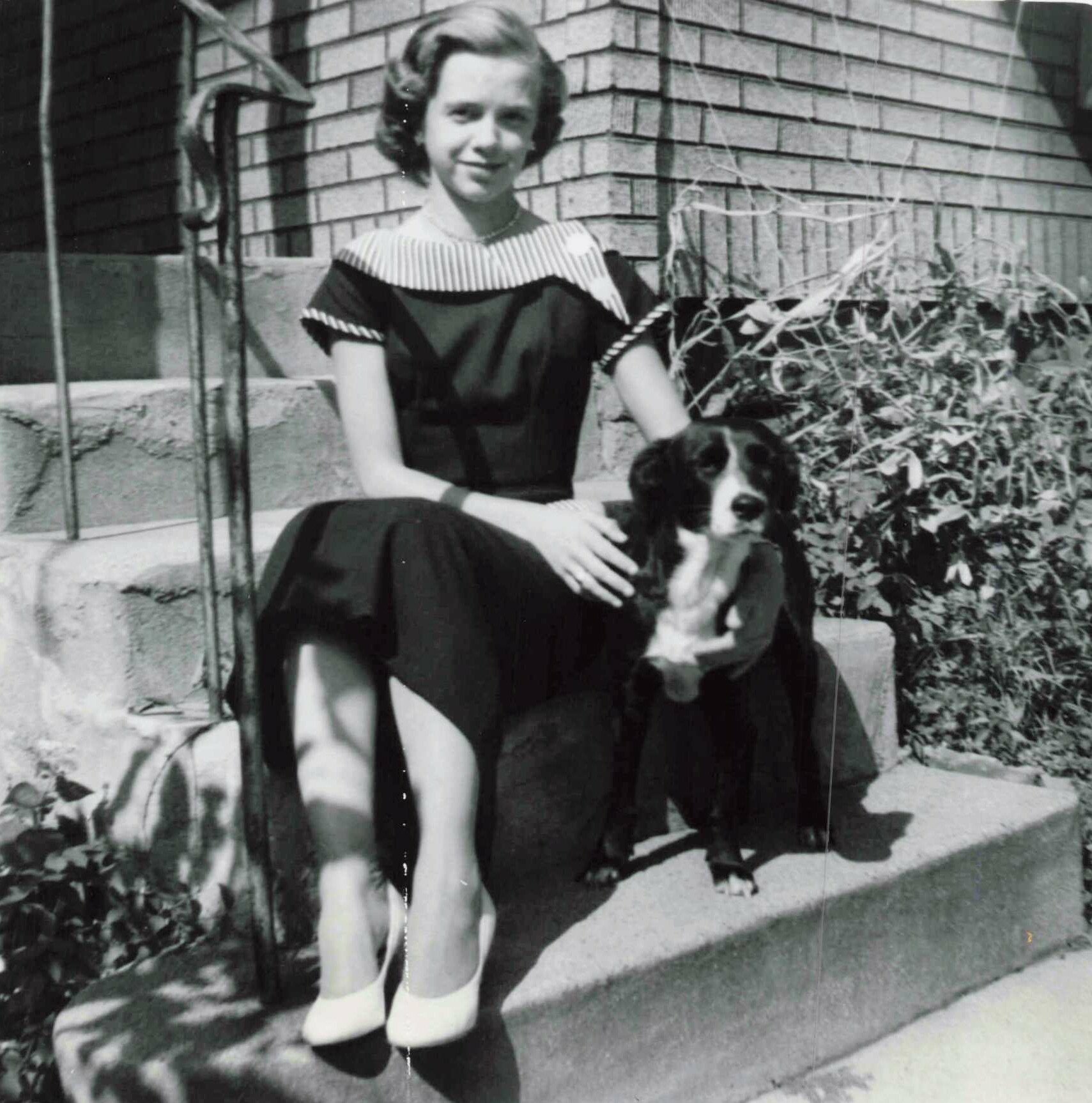 Young woman in a vintage dress sits on concrete steps with a black-and-white spaniel beside her, in front of a brick wall of a house.