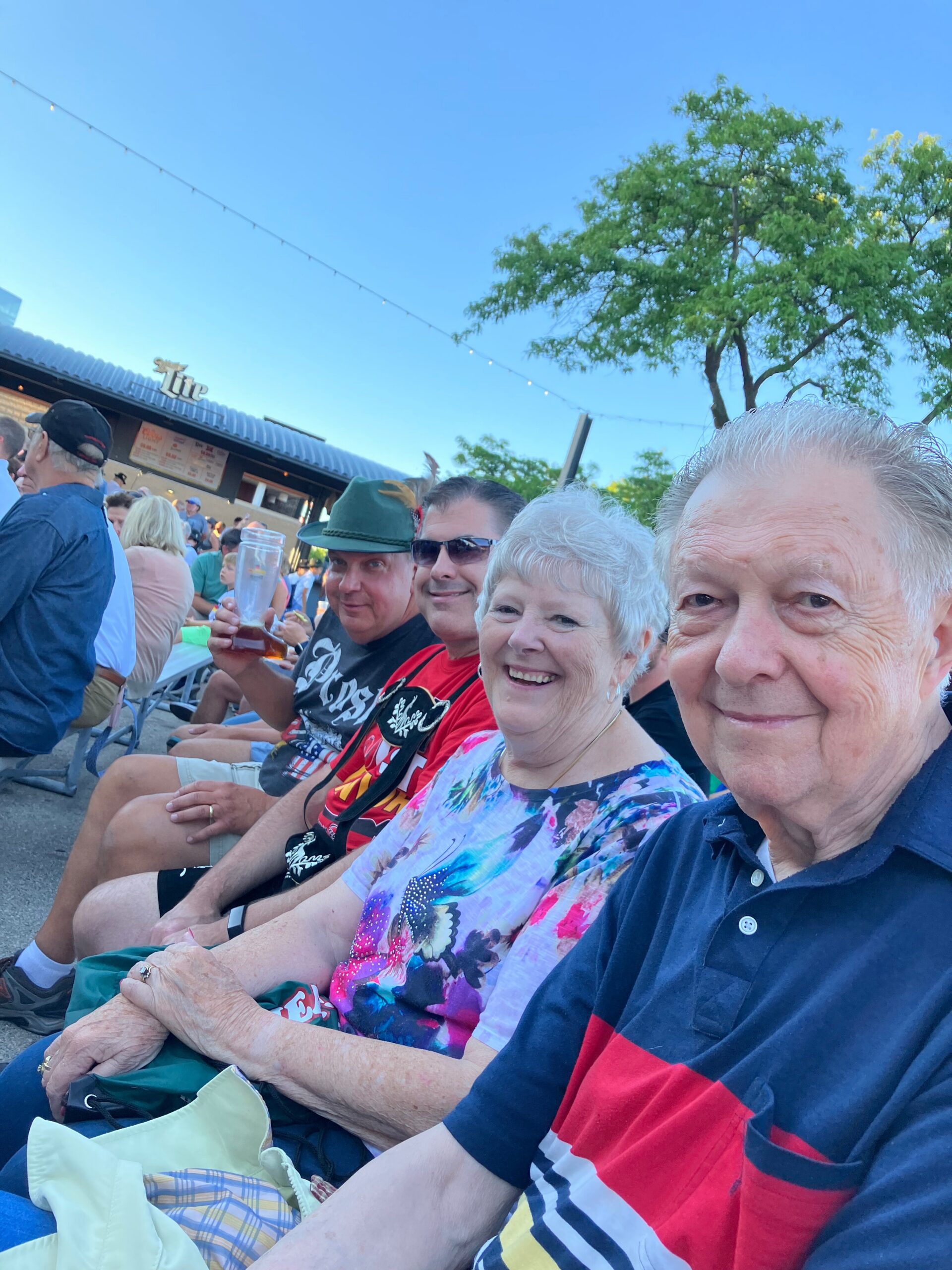 Group of smiling seniors seated at an outdoor event, long line of people at a food stand in the background, blue sky above.