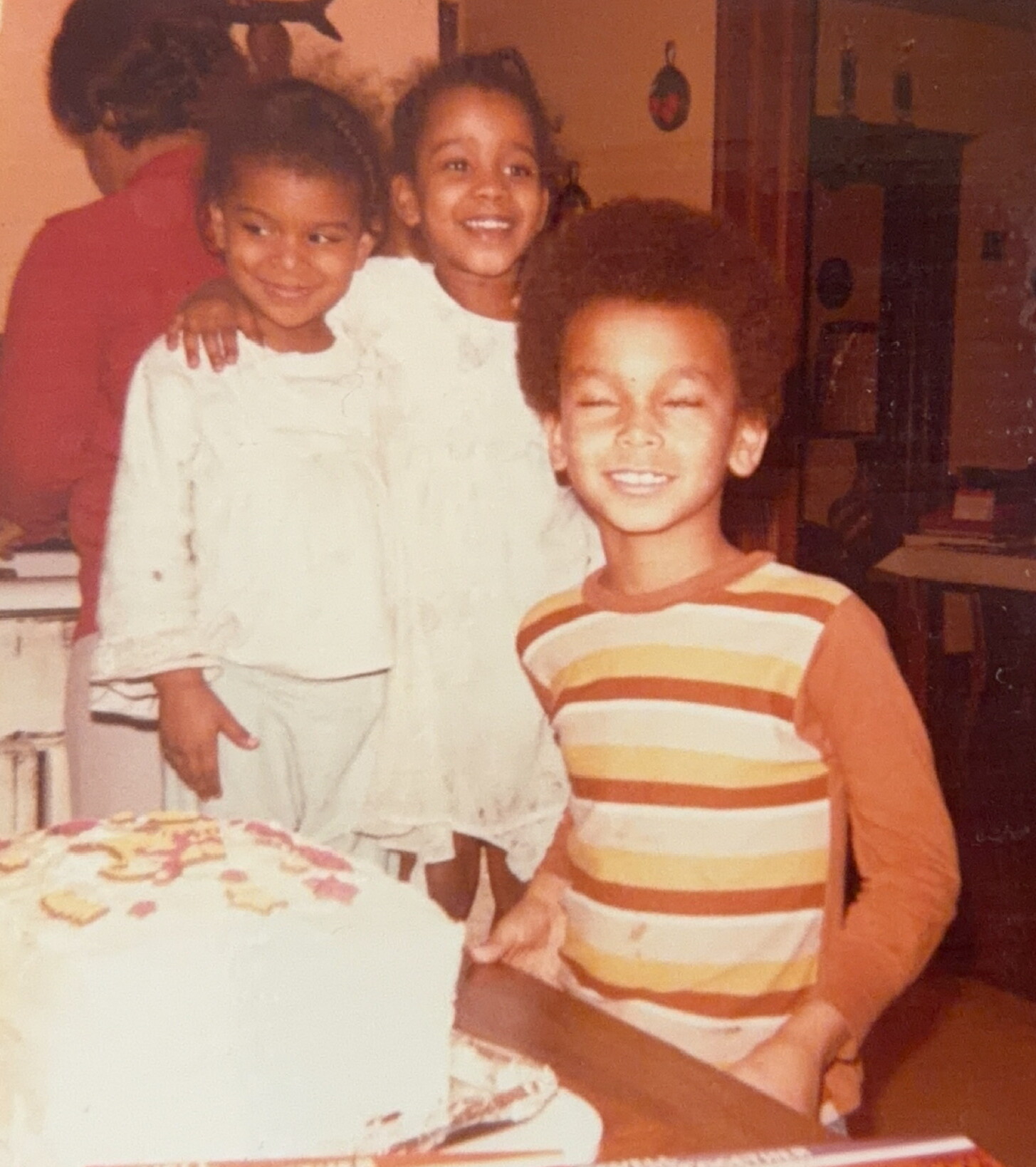 Three children stand together smiling beside a birthday cake on a table indoors at a party.
