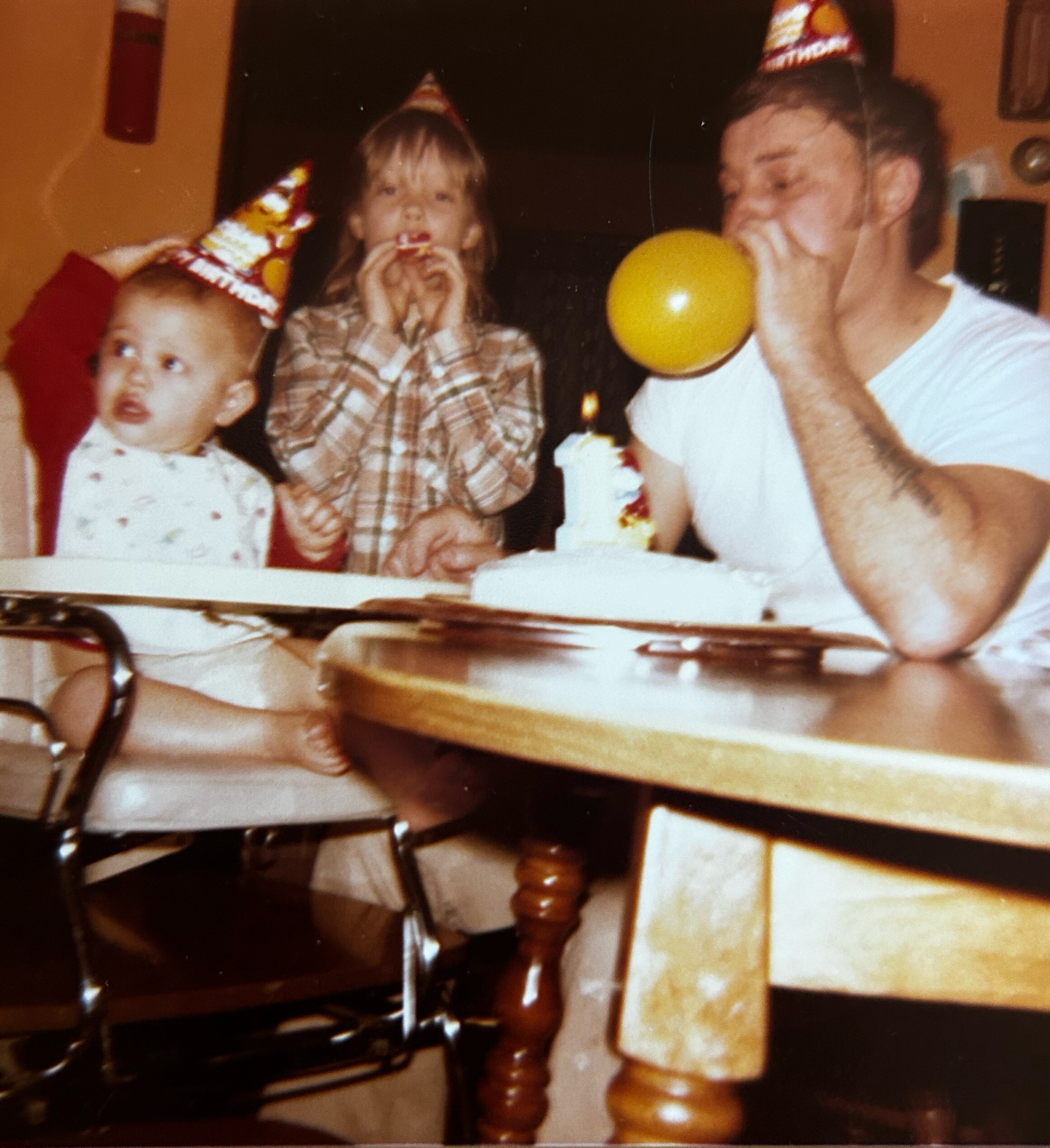 Three people at a birthday party: a man inflates a yellow balloon while two children wearing party hats watch nearby.