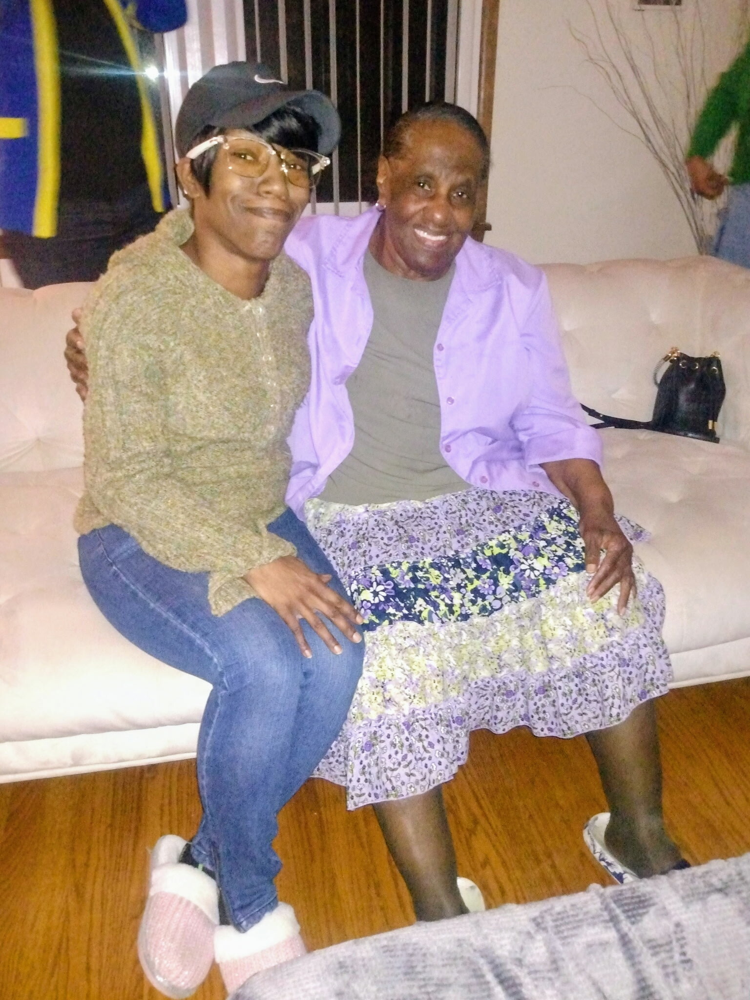 Two women sit closely on a beige couch, smiling at the camera in a living room setting, one wearing a hat and glasses while the other wears a lavender shirt.