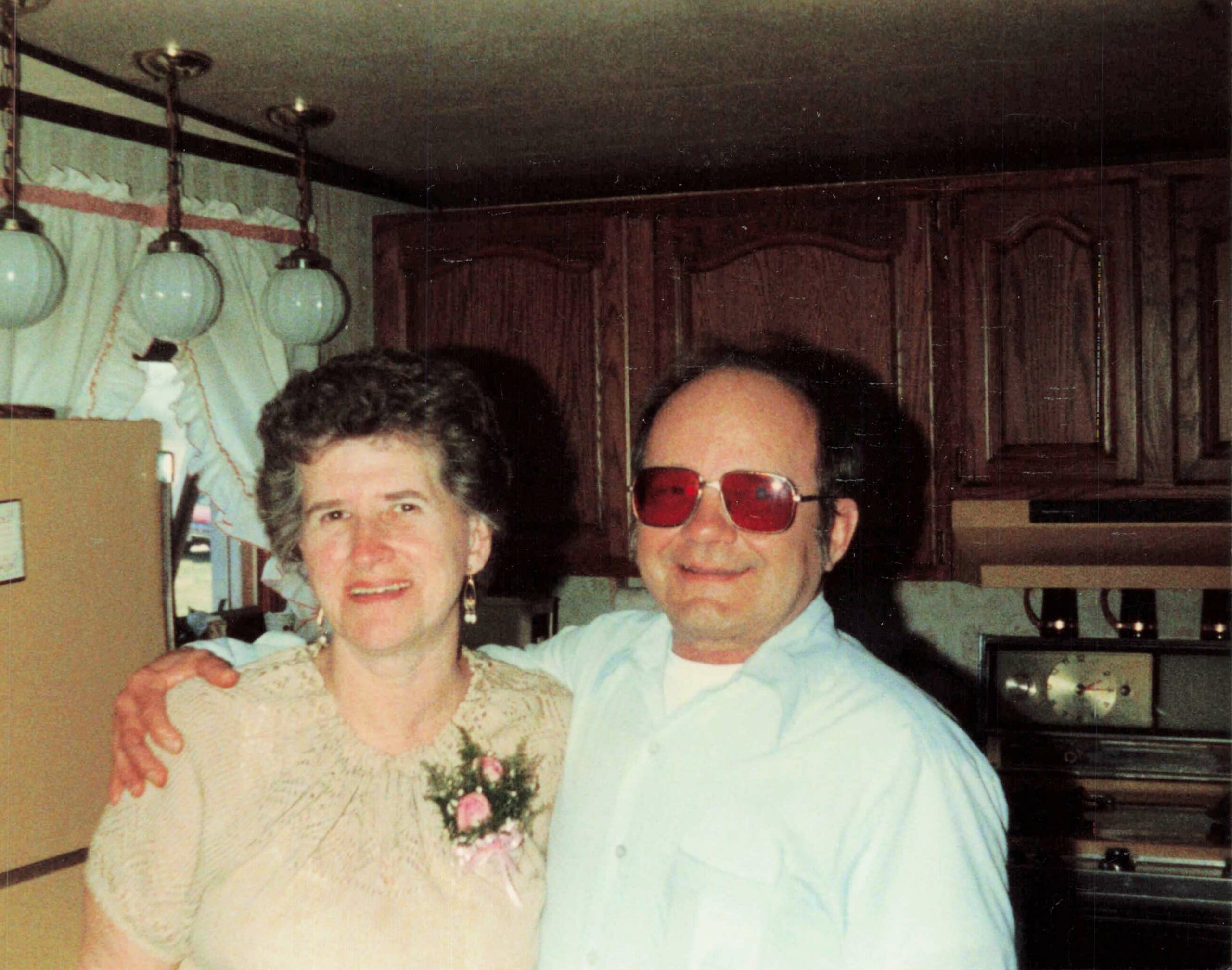 An elderly couple poses together in a kitchen, the man wearing red-tinted glasses and a light blue shirt, his arm around the woman in a floral blouse.