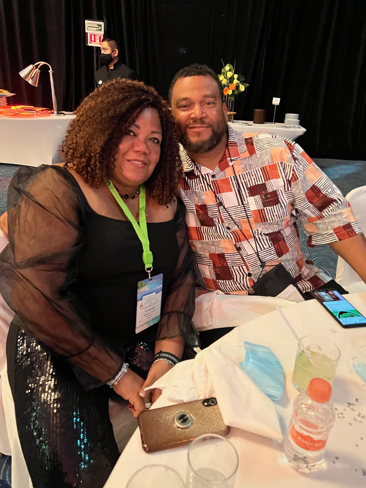 Two smiling guests sit side by side at a banquet table; woman with a green lanyard and a man in a geometric orange–brown shirt pose for a photo, with drinks and a phone on the table and a buffet area in the background.
