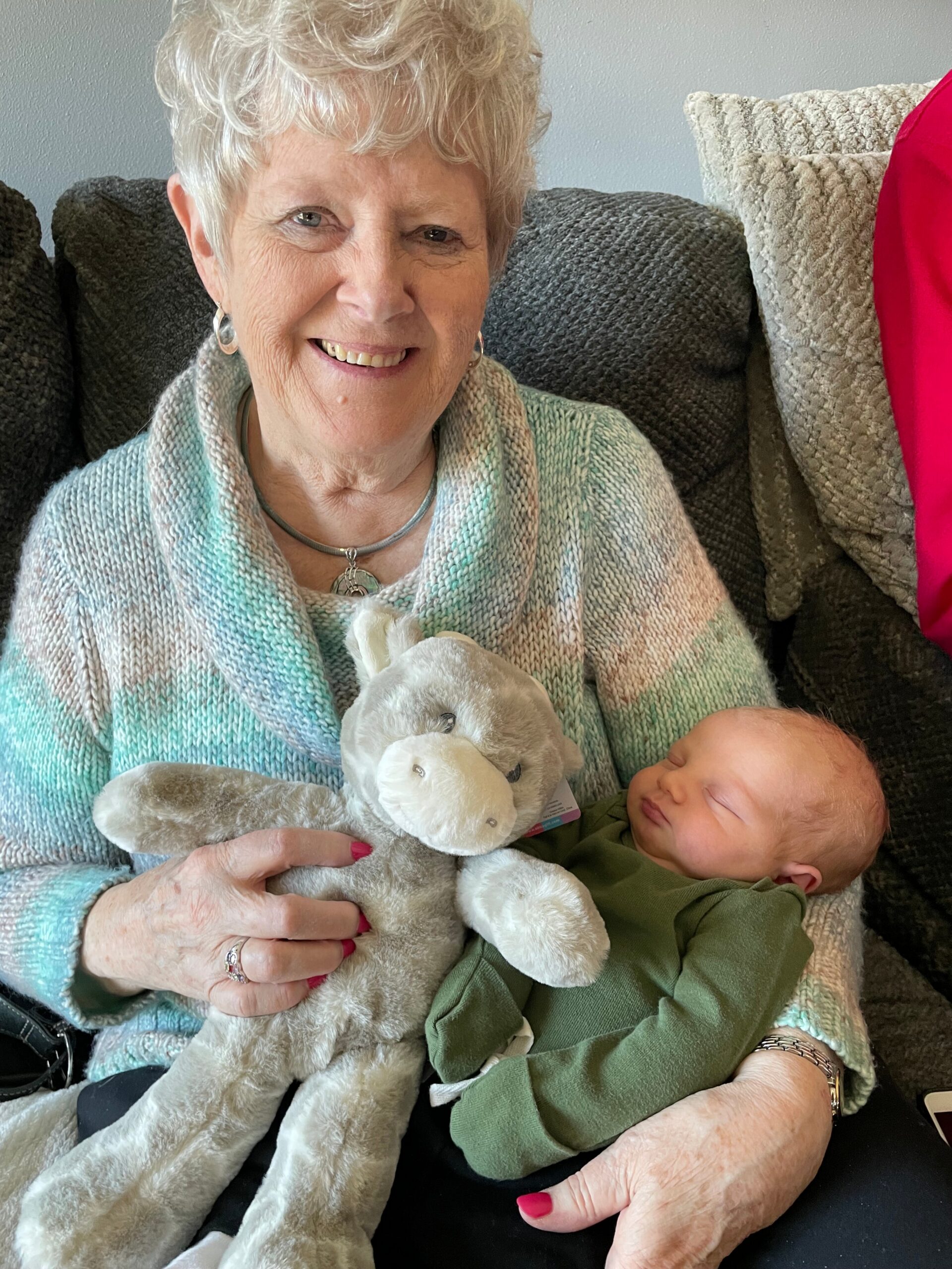 Smiling elderly woman with short white hair sits on a sofa, holding a plush bunny while a newborn sleeps on her lap.