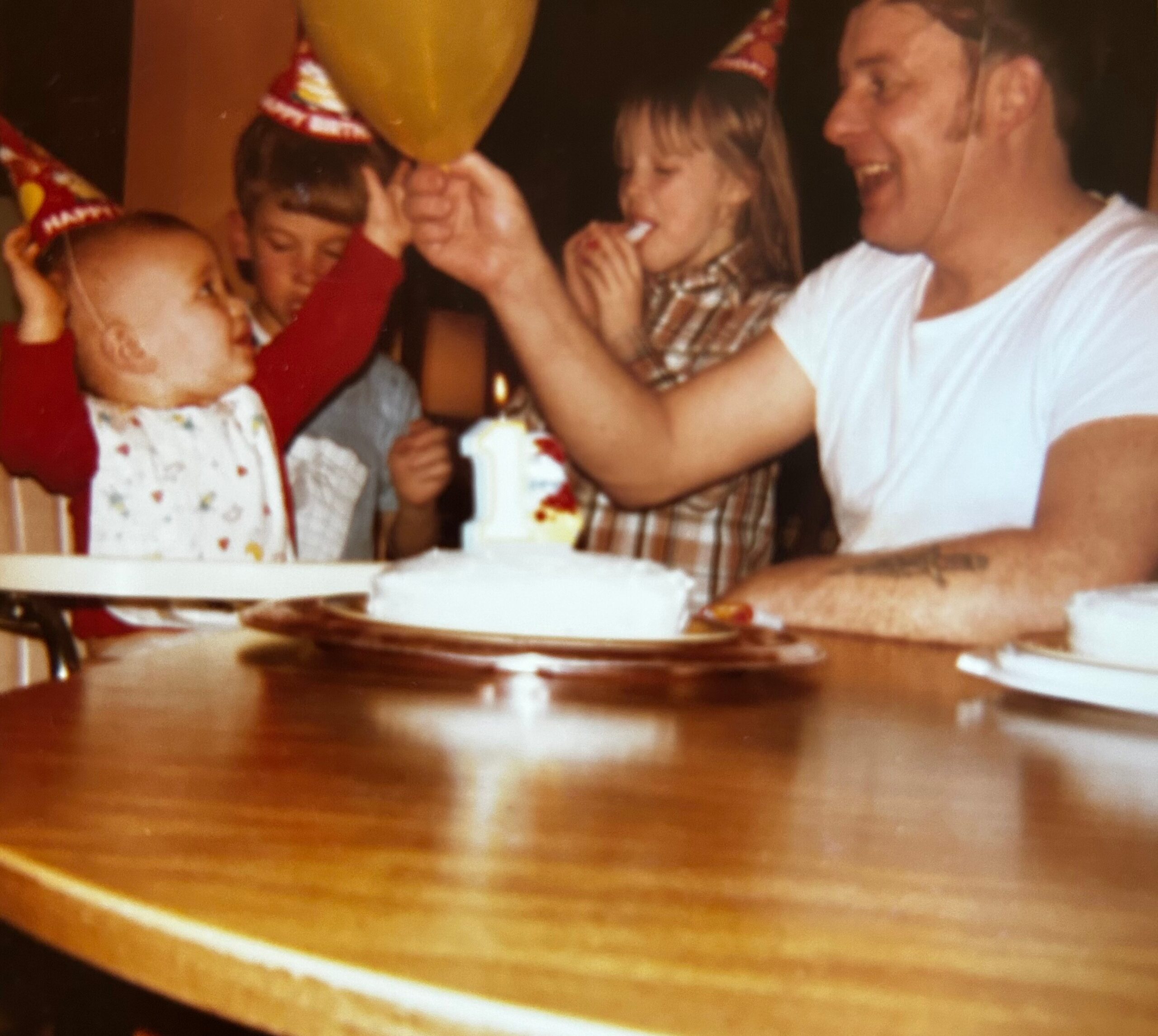 Children wearing party hats gather around a birthday cake with lit candles at a celebration table.