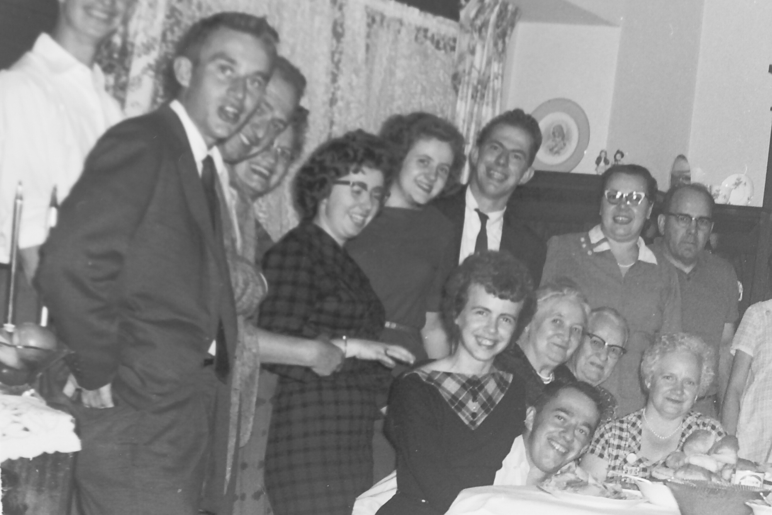 Black-and-white photo of a large group of adults at a party, smiling and posing around a table with a decorated cake.