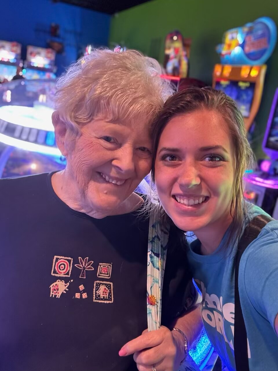Two women smiling for a selfie in a colorful arcade with arcade cabinets in the background, neon lights glowing around them.