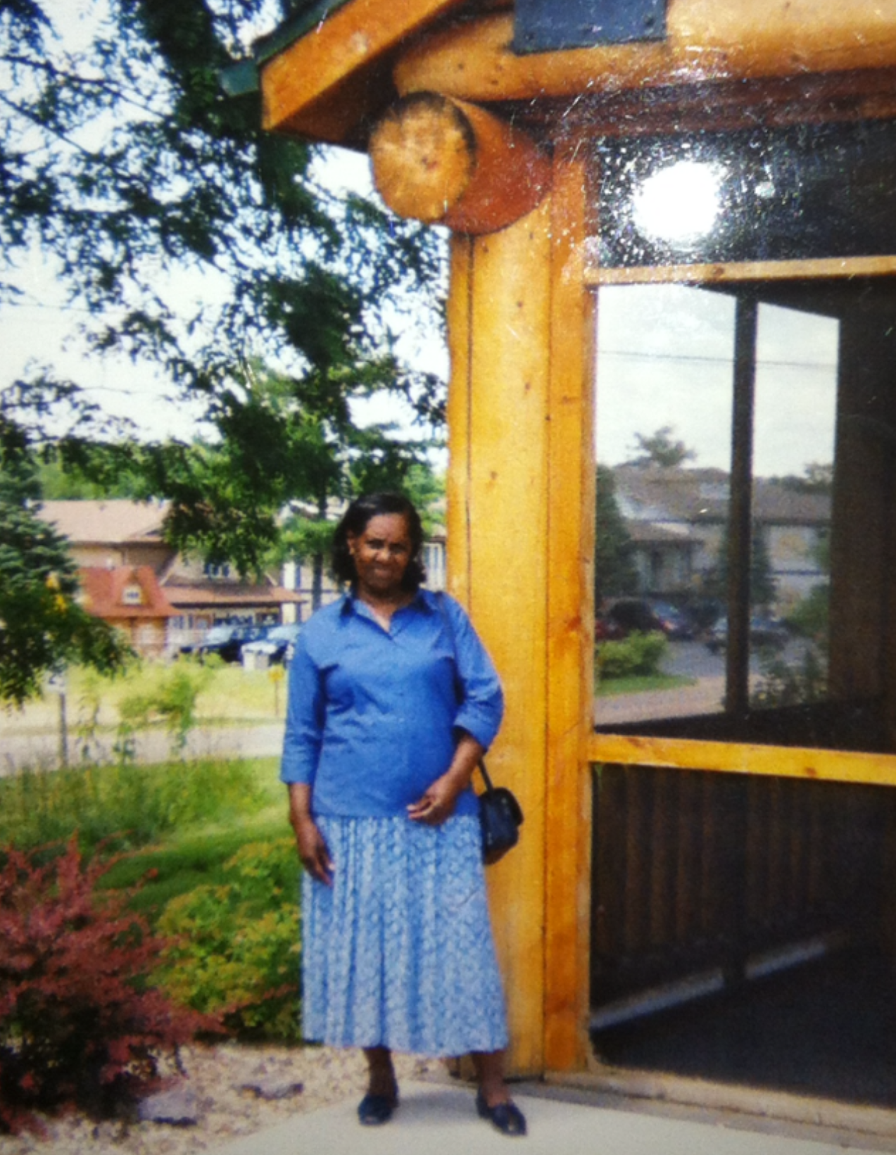 Woman in blue blouse and patterned skirt standing outside a wooden building with large windows and greenery in the background.