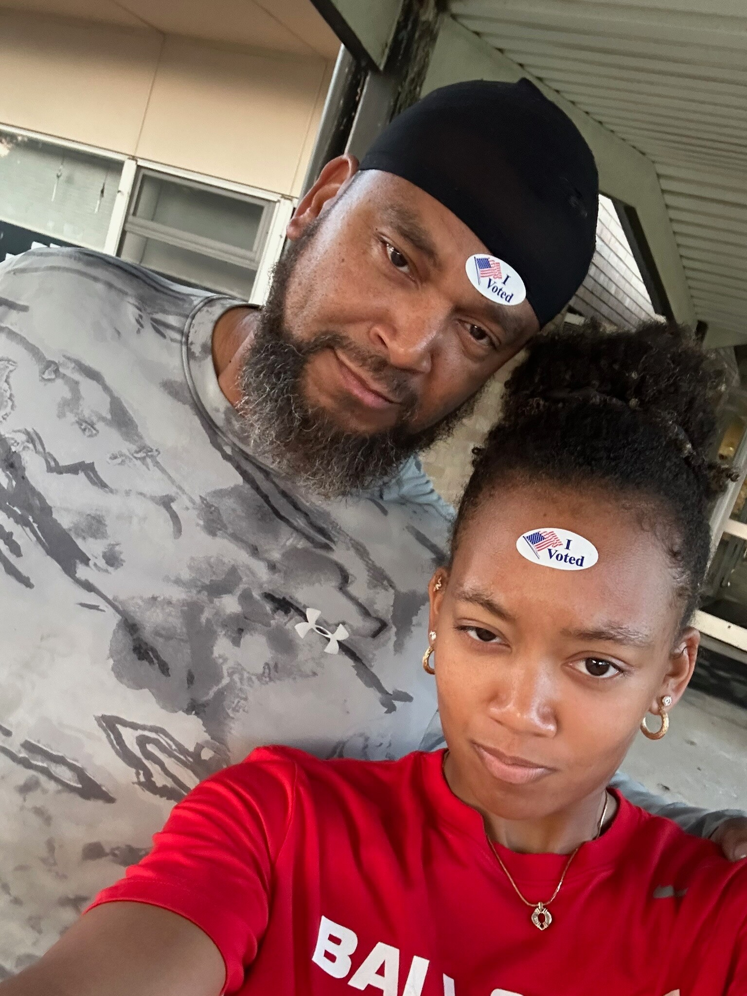 Two people take a close-up selfie outdoors, a man with a black durag and beard beside a woman, both wearing 'I Voted' stickers on their foreheads/surfaces.