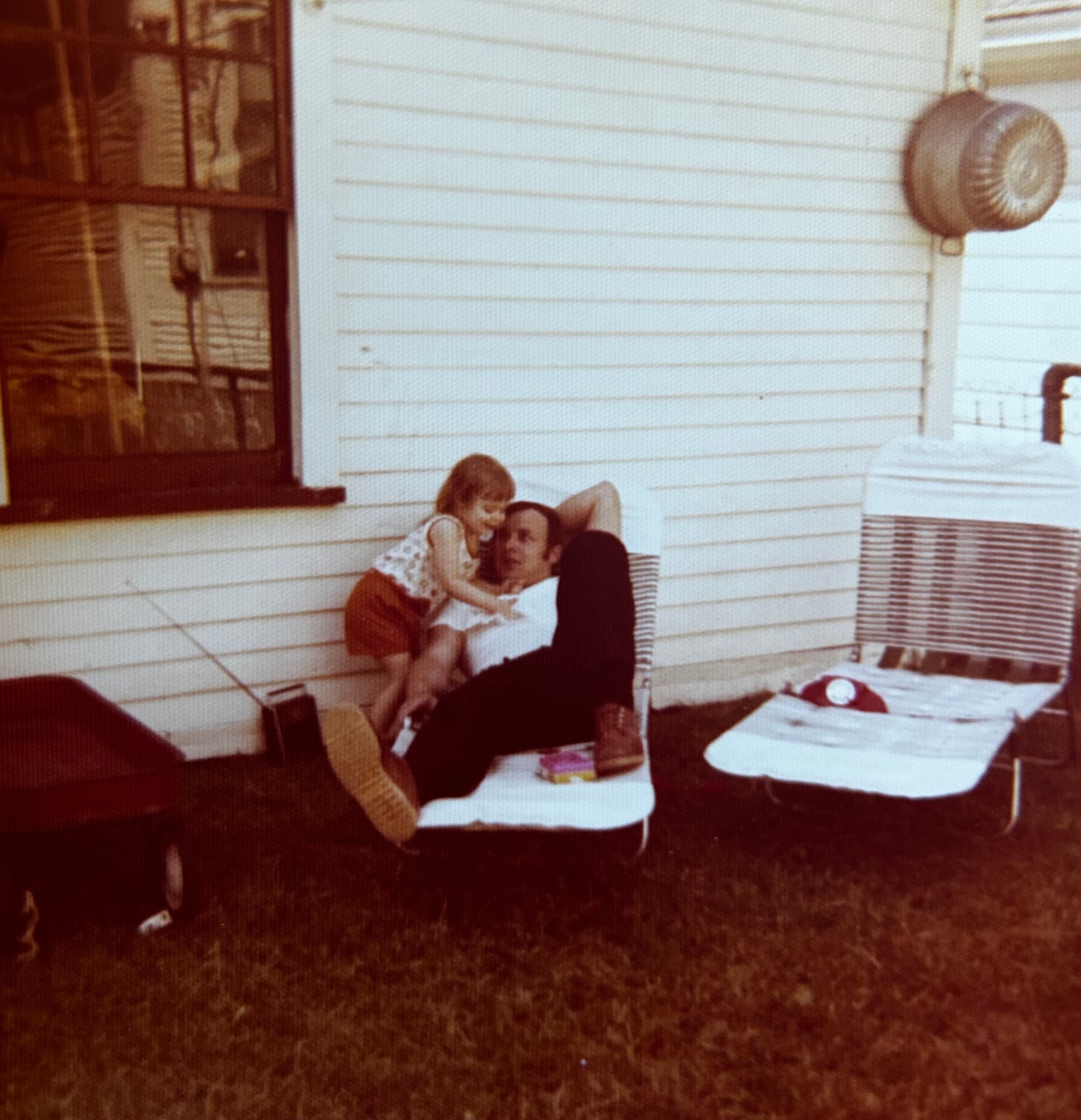 Children sitting on white steps outside a house, leaning together and smiling at the camera near a large outdoor faucet/vent area.