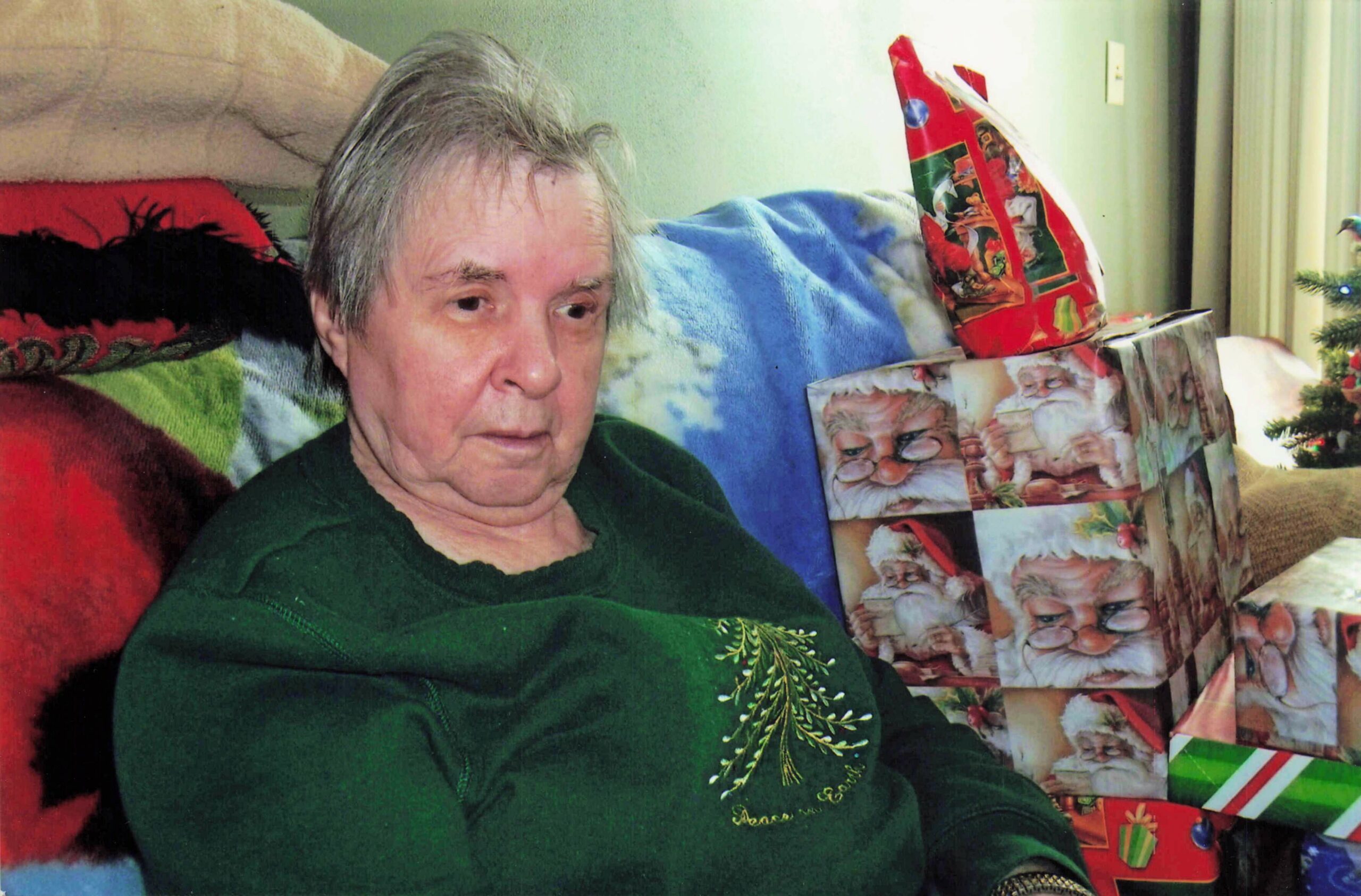 Older man with gray hair wearing a green sweater sits on a couch surrounded by wrapped Christmas gifts and blankets in a living room.