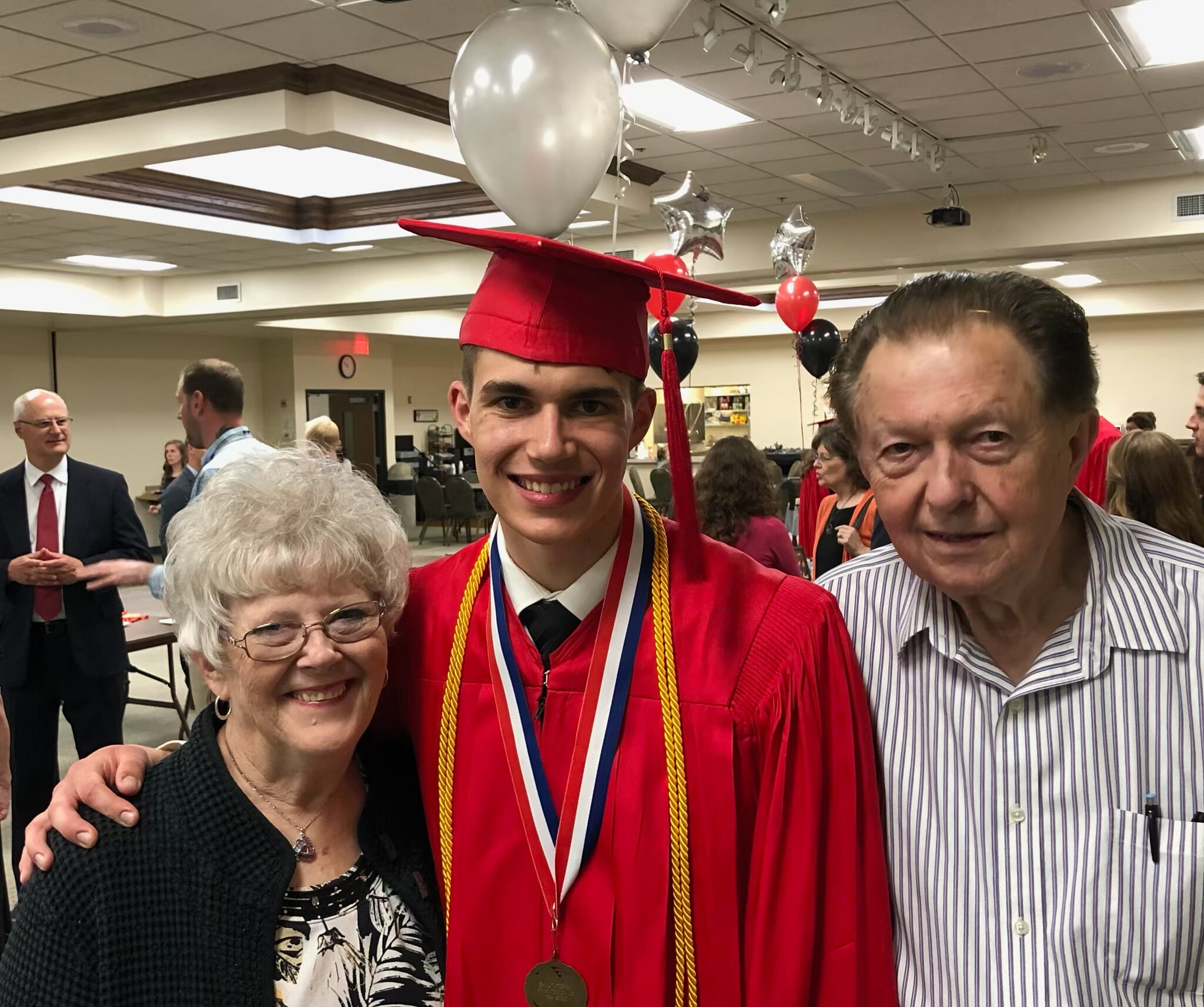 Young graduate in a red cap and gown smiling at a celebration, wearing medals and cords indoors among attendees.