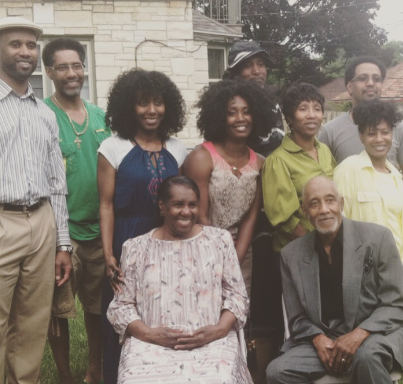 Nine adults posing together outdoors in front of a stone house, some standing and two seniors seated in front.