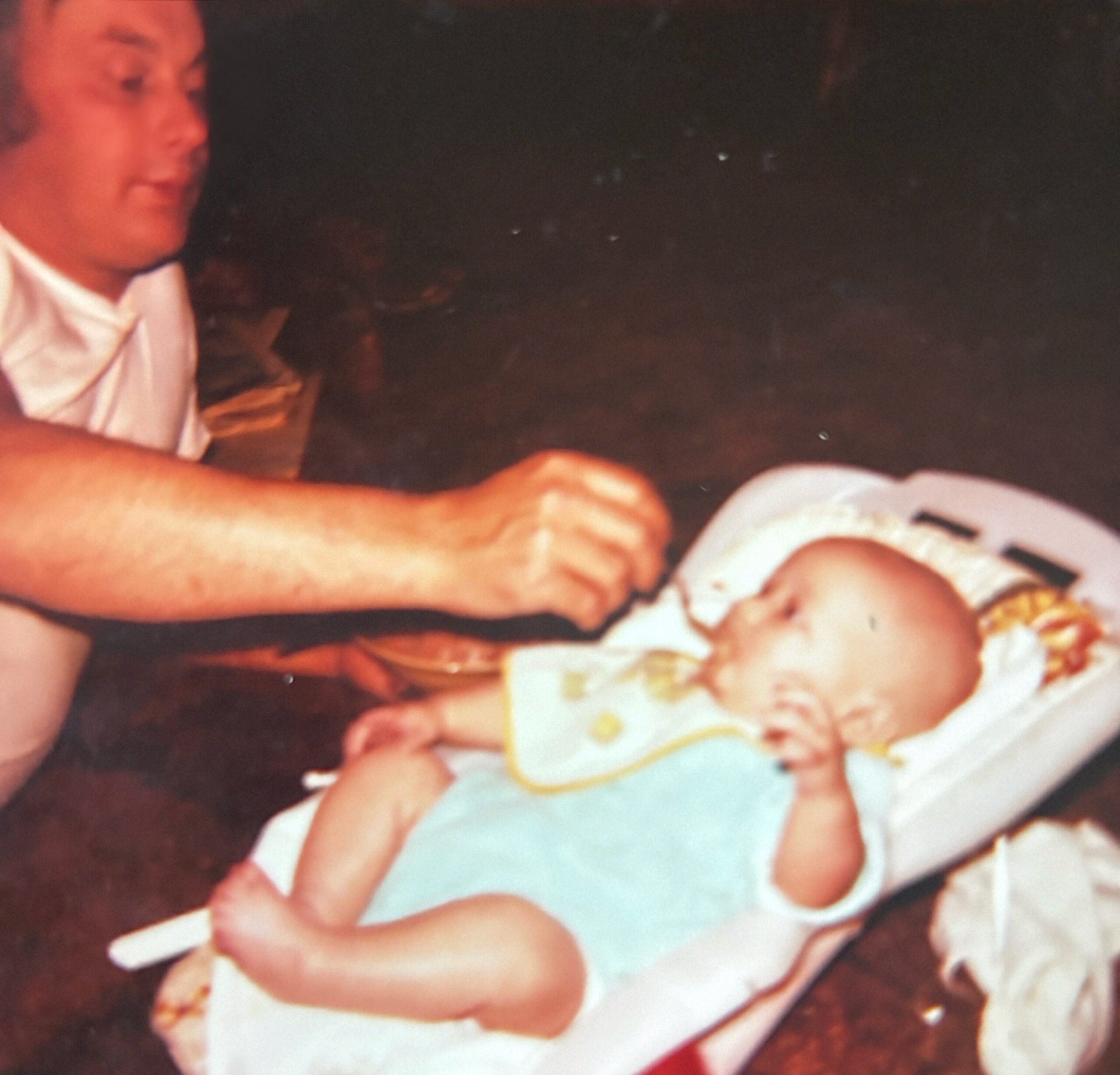 Close-up of a baby lying on a blanket, wearing a diaper, with an adult nearby in a dimly lit room.