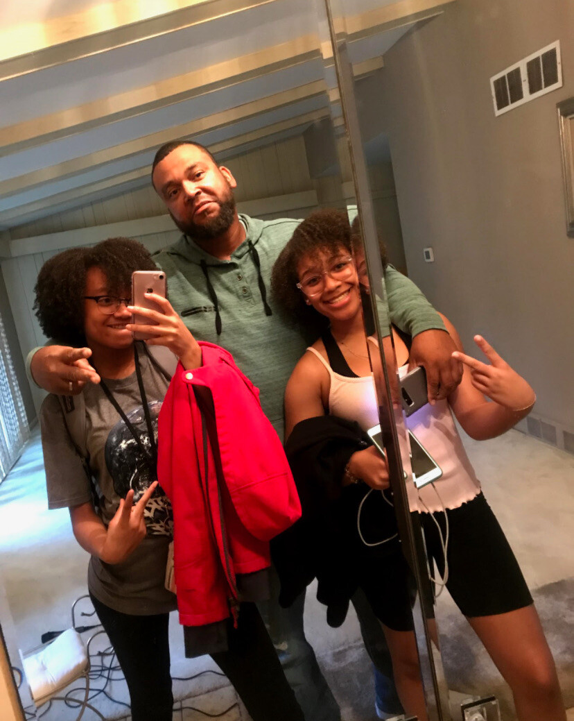 Three people pose for a mirror selfie: a man in a green hoodie with two girls on either side, all smiling.