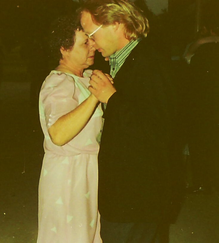 Older woman in a pale pink dress dancing closely with a man in a dark suit, their foreheads touching as they hold hands.