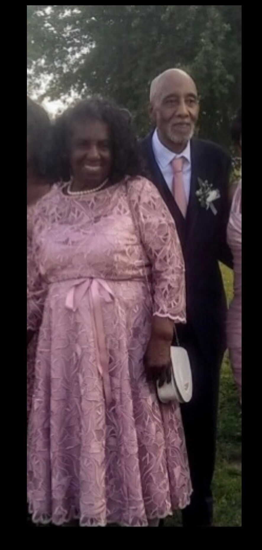 Couple at a formal outdoor event: woman in a pink lace dress with belt beside a man in a suit and tie with a boutonniere.