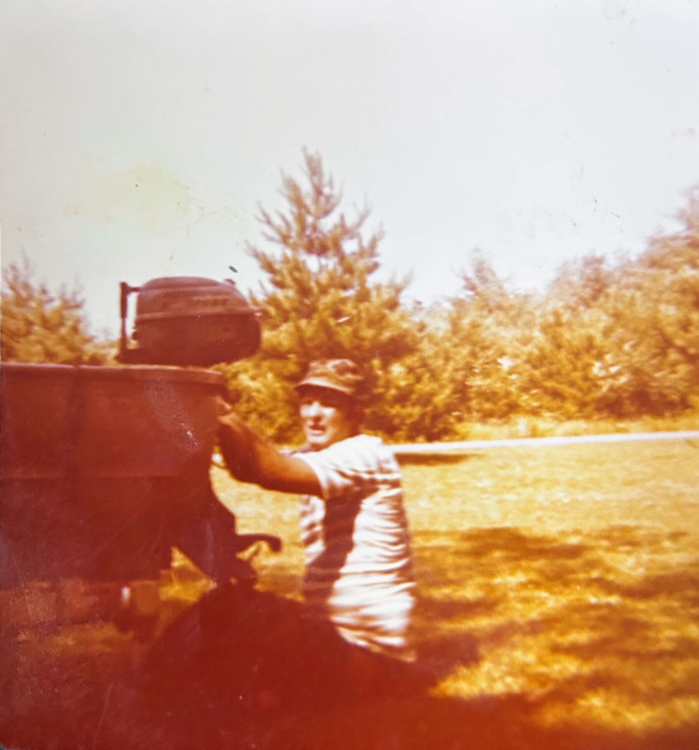 Person in a light-colored shirt stands beside a large dark vehicle or object outdoors, with trees in the background and a warm, vintage color cast.
